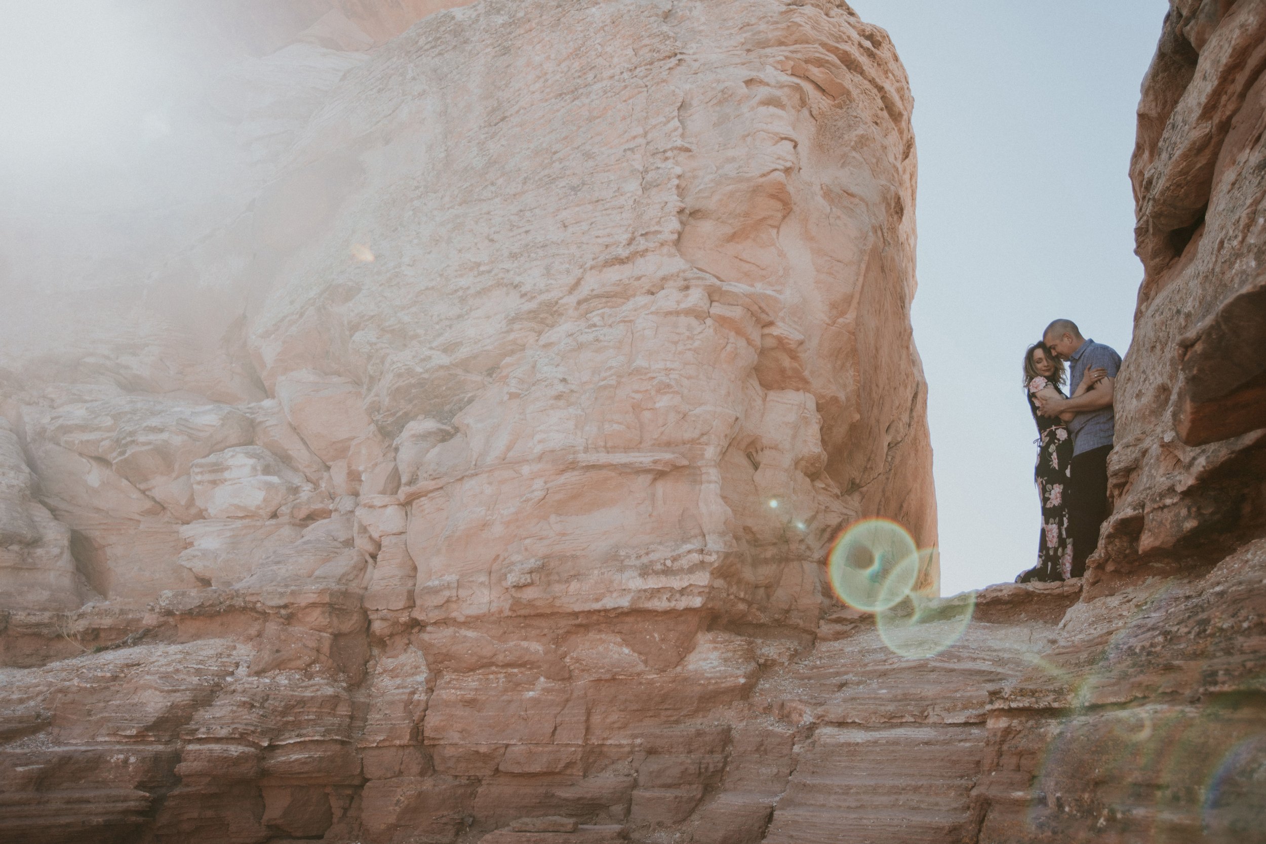 Couple embracing on the edge of a desert canyon, with a sunflare adding a dramatic and adventurous touch to the scene.