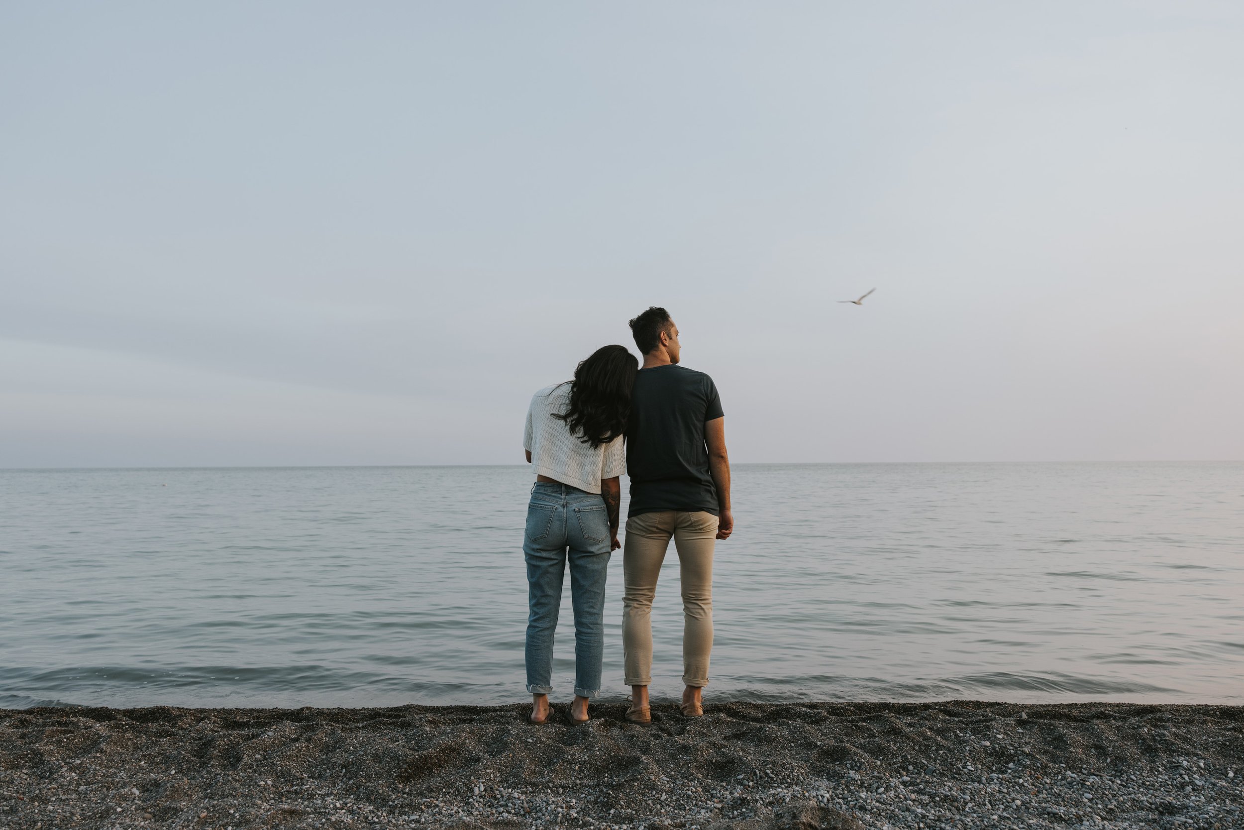 A couple stands side by side on a beach, gazing out at the horizon as the sun sets, their relaxed connection and the serene atmosphere captured beautifully.