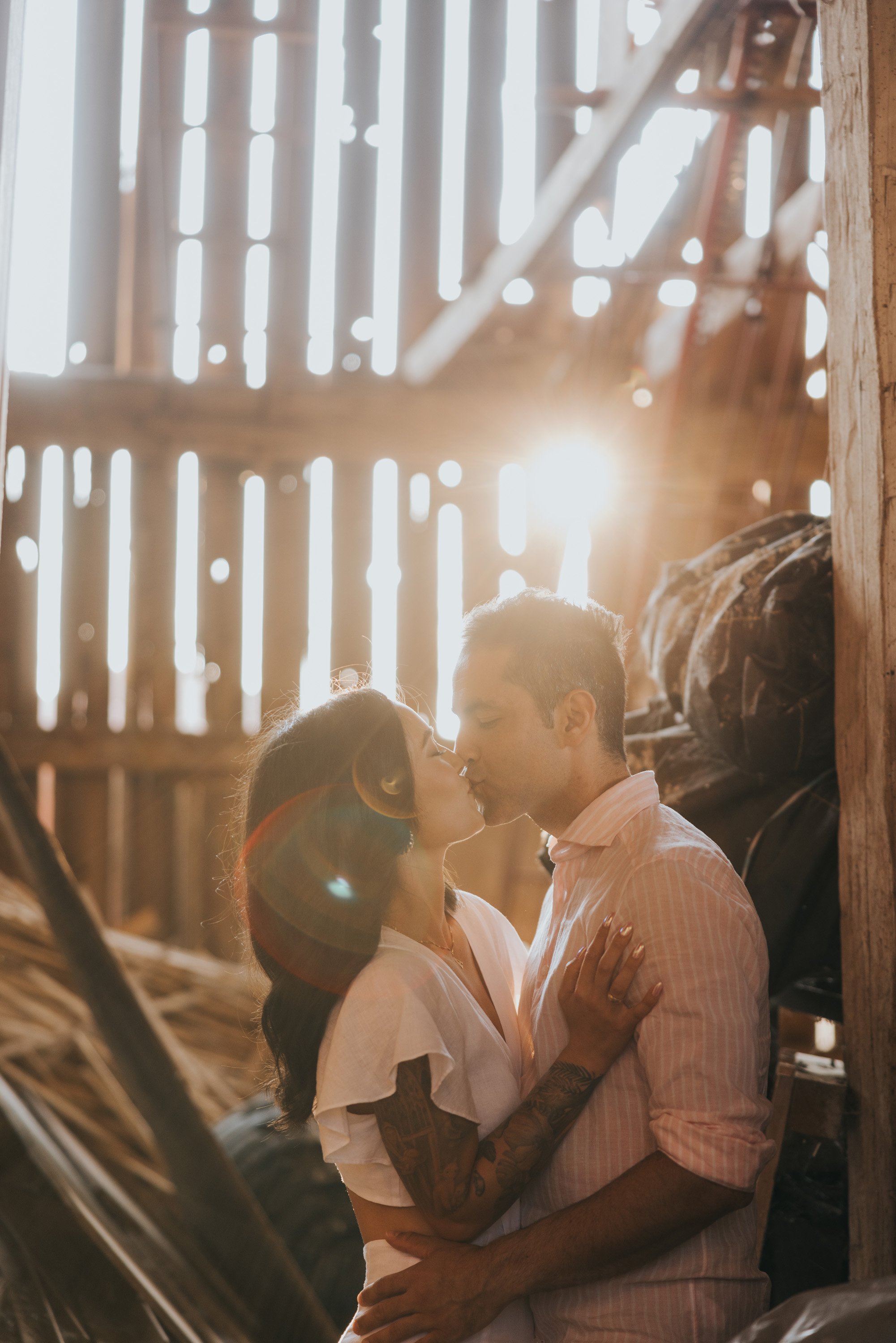 A couple sharing a kiss inside a rustic barn as the setting sun casts a warm glow through the wooden slats.