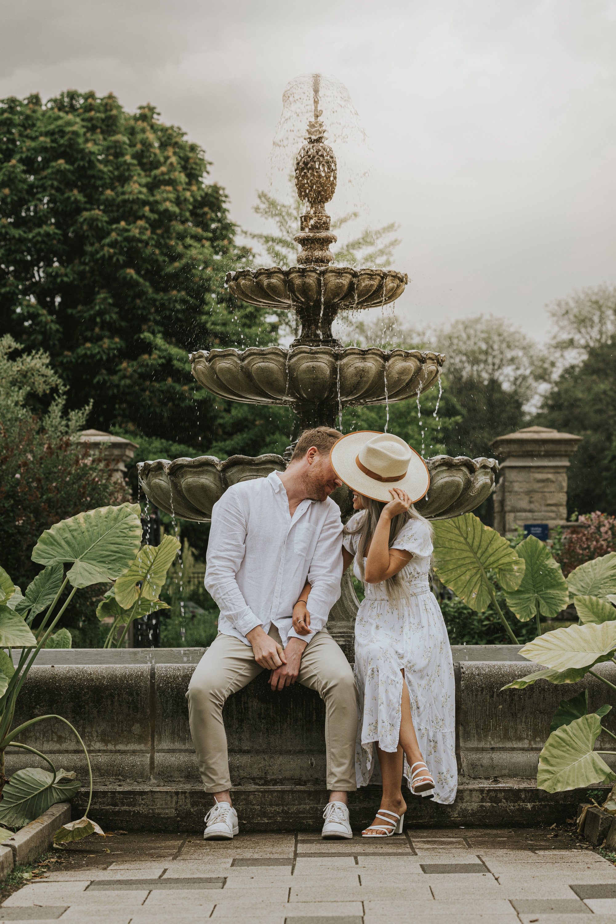 A couple shares a laugh while sitting by a fountain in a romantic garden, with one partner playfully covering their face with a hat.