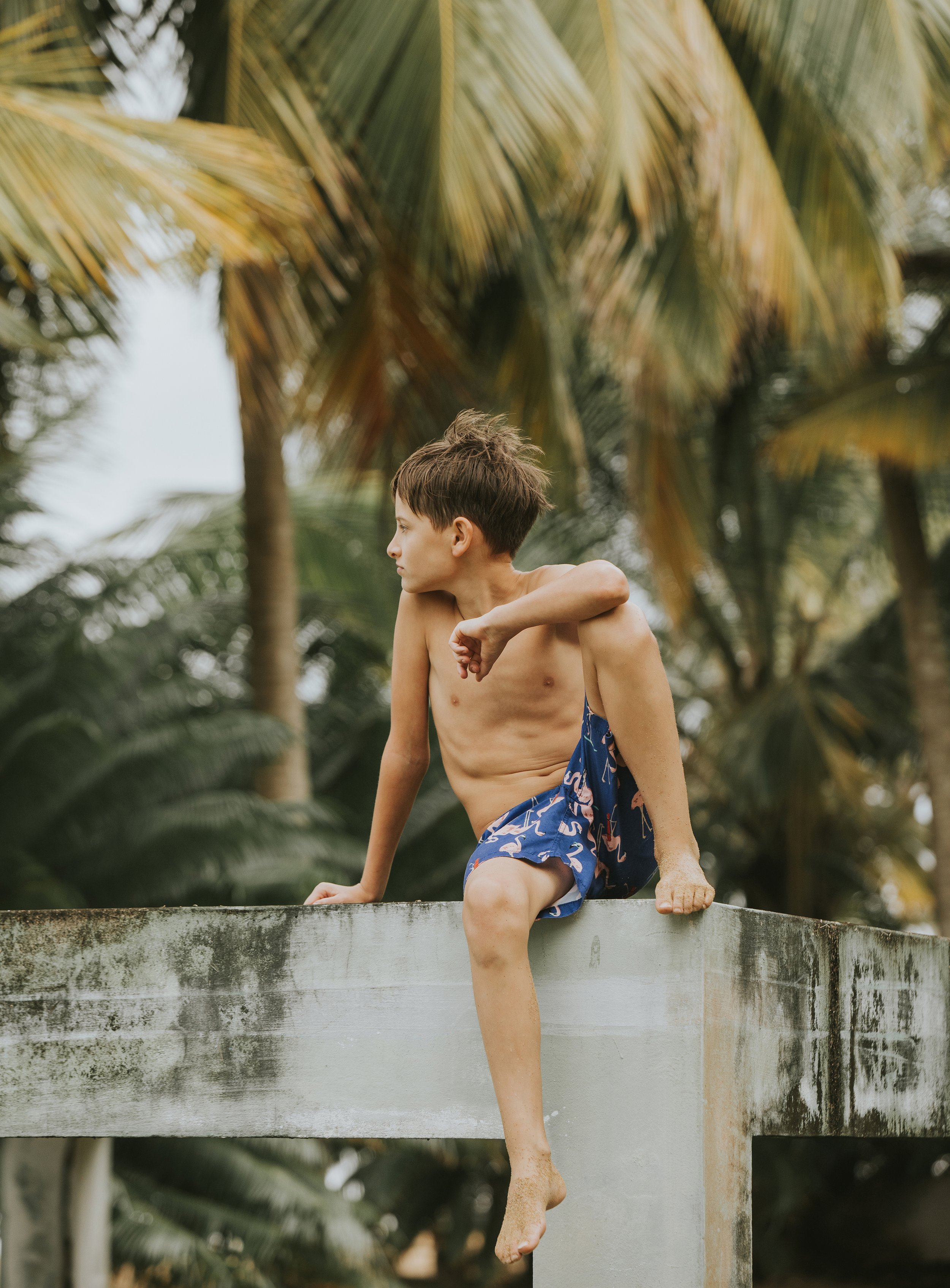 A boy in swim shorts sitting on a concrete ledge with tropical palm trees in the background.