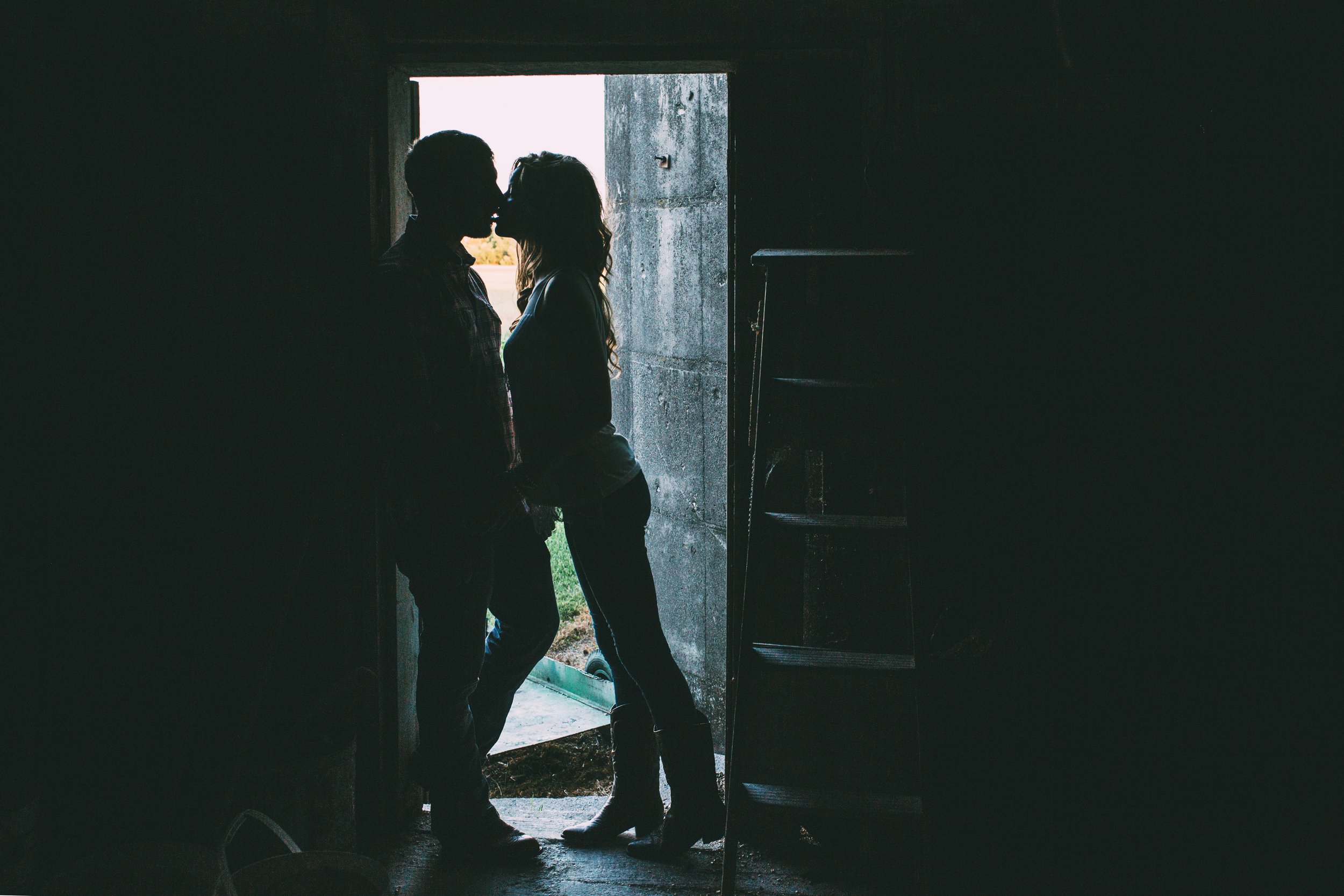 A couple shares a romantic kiss in the silhouette of a farm doorway during sunset, framed by the rustic charm of the setting, evoking feelings of warmth and love.