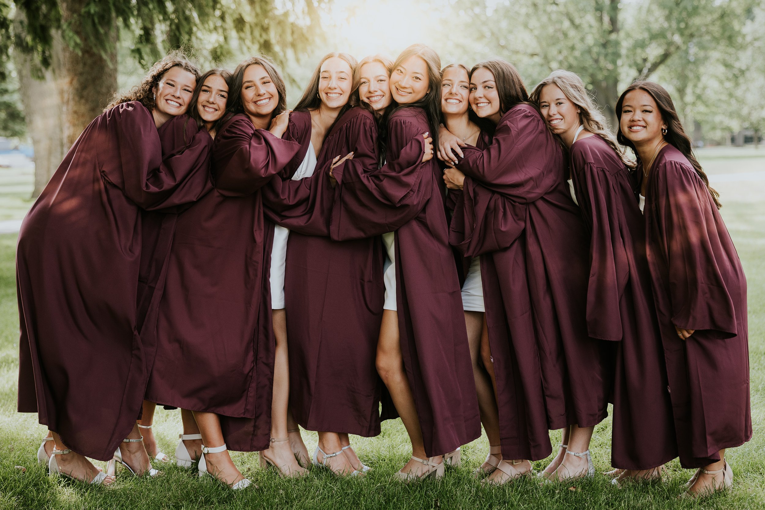 A group of graduates in maroon caps and gowns hug tightly, celebrating together outdoors.
