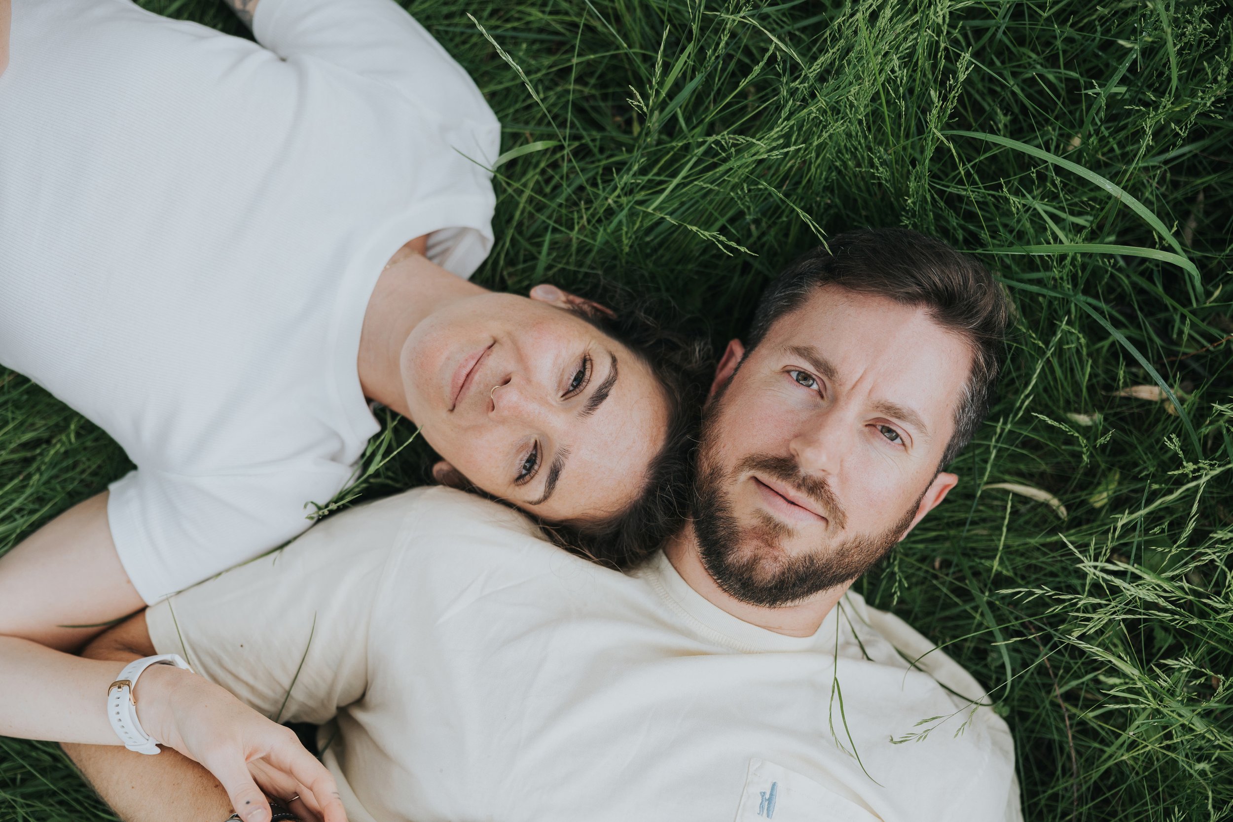 A couple lies in tall grass, sharing a peaceful and loving moment, with a rustic, natural setting enhancing their connection.