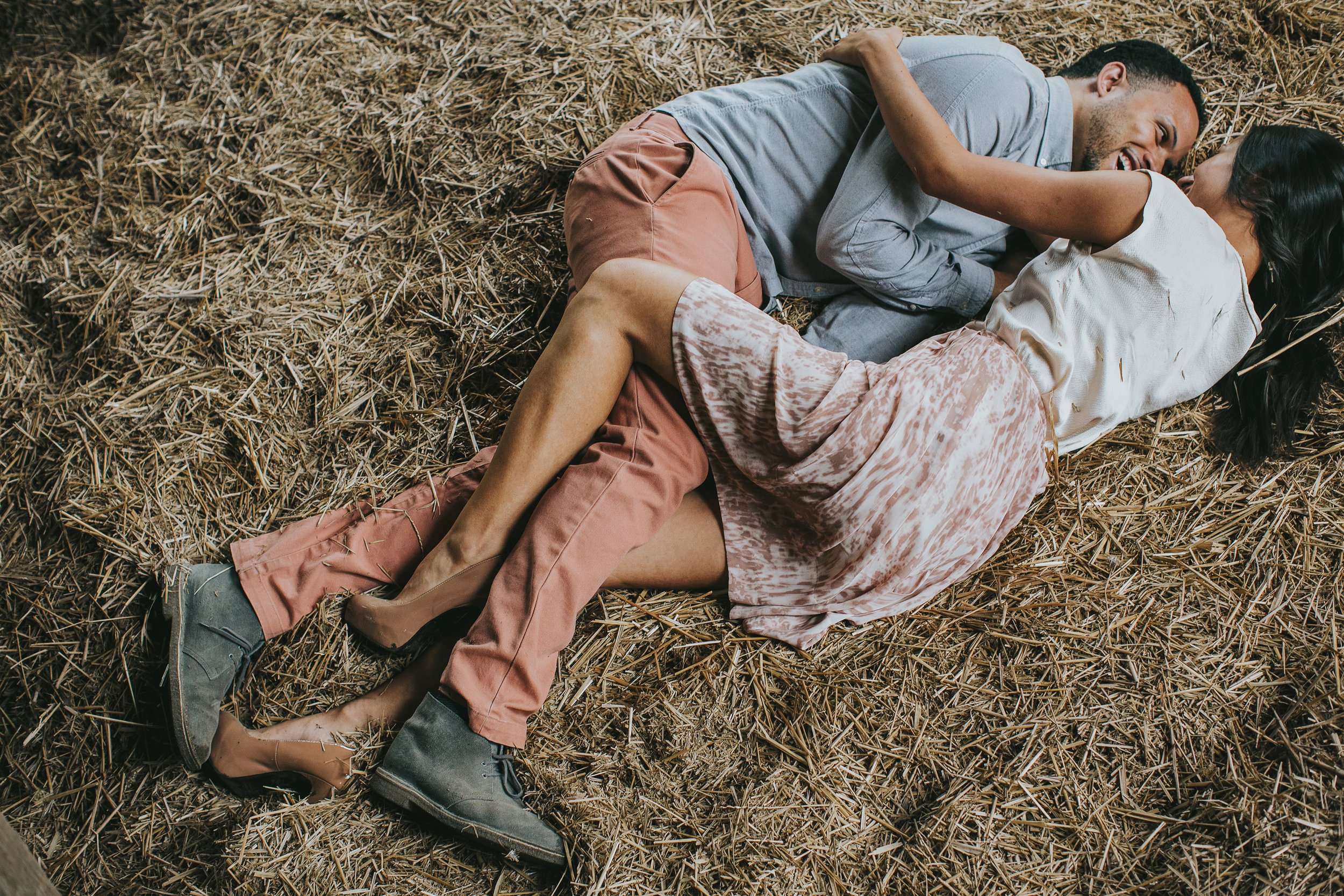 A couple lying close together on hay inside a rustic barn, capturing an intimate and playful moment during their engagement.