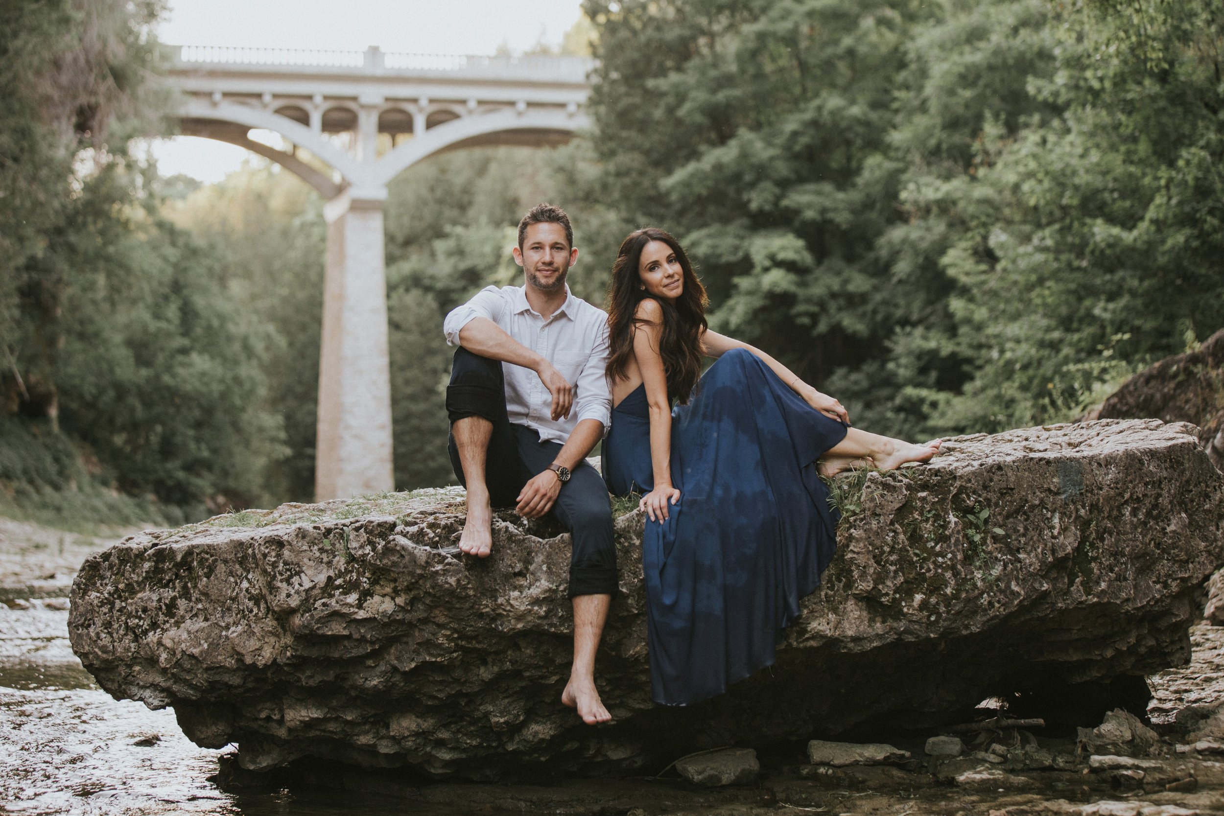 A couple sits barefoot on a large rock by the Elora Gorge, with a picturesque bridge in the background, enjoying a relaxed moment in nature.