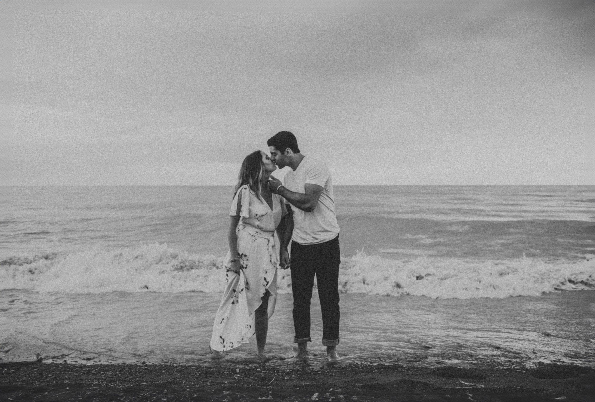 A black-and-white photo captures a couple kissing by the waves on a beach, their intimate moment framed by the natural beauty of the ocean.