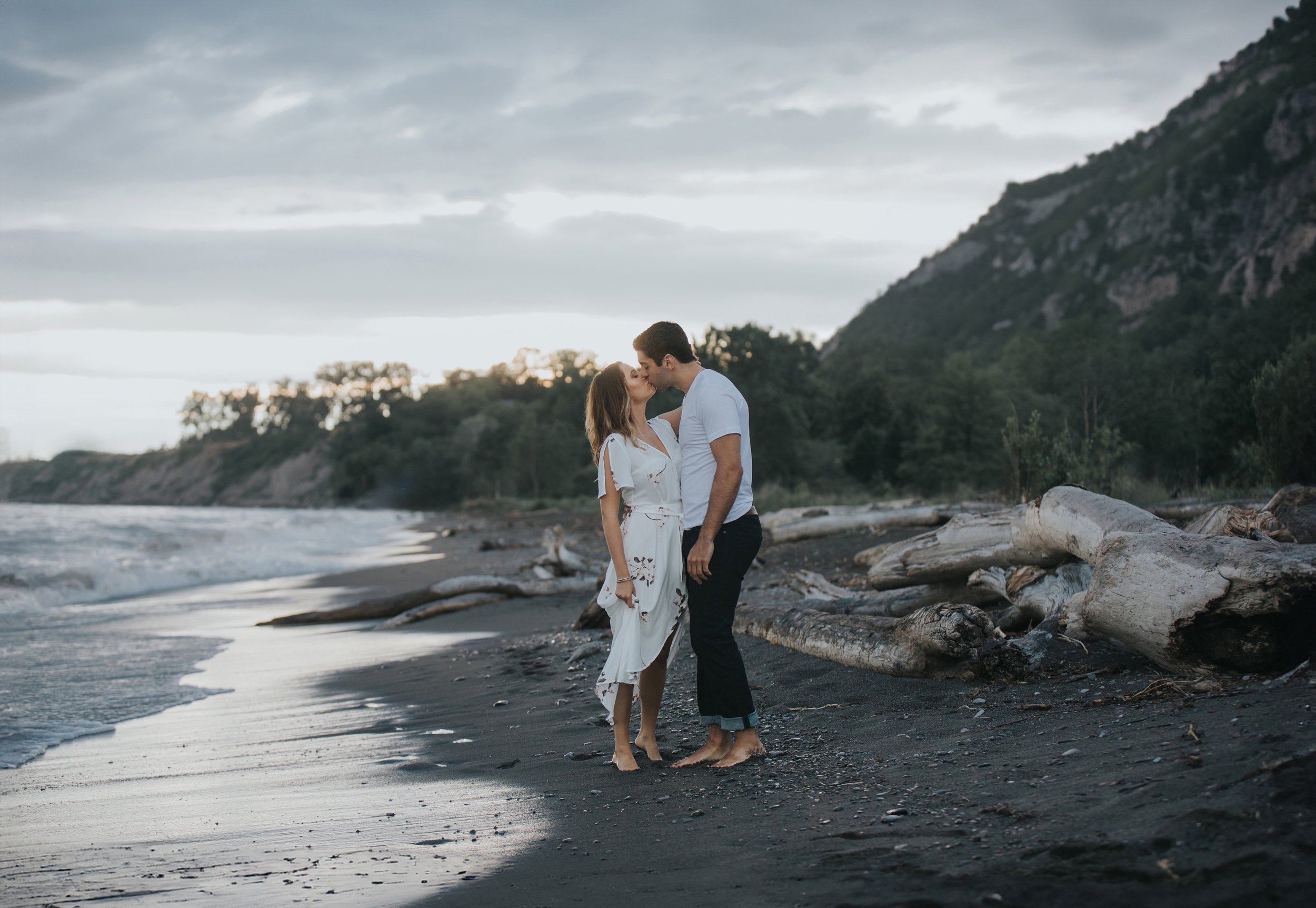 A couple shares a romantic kiss on a driftwood-strewn beach with mountains in the background. The dramatic scenery enhances the intimate connection between them.