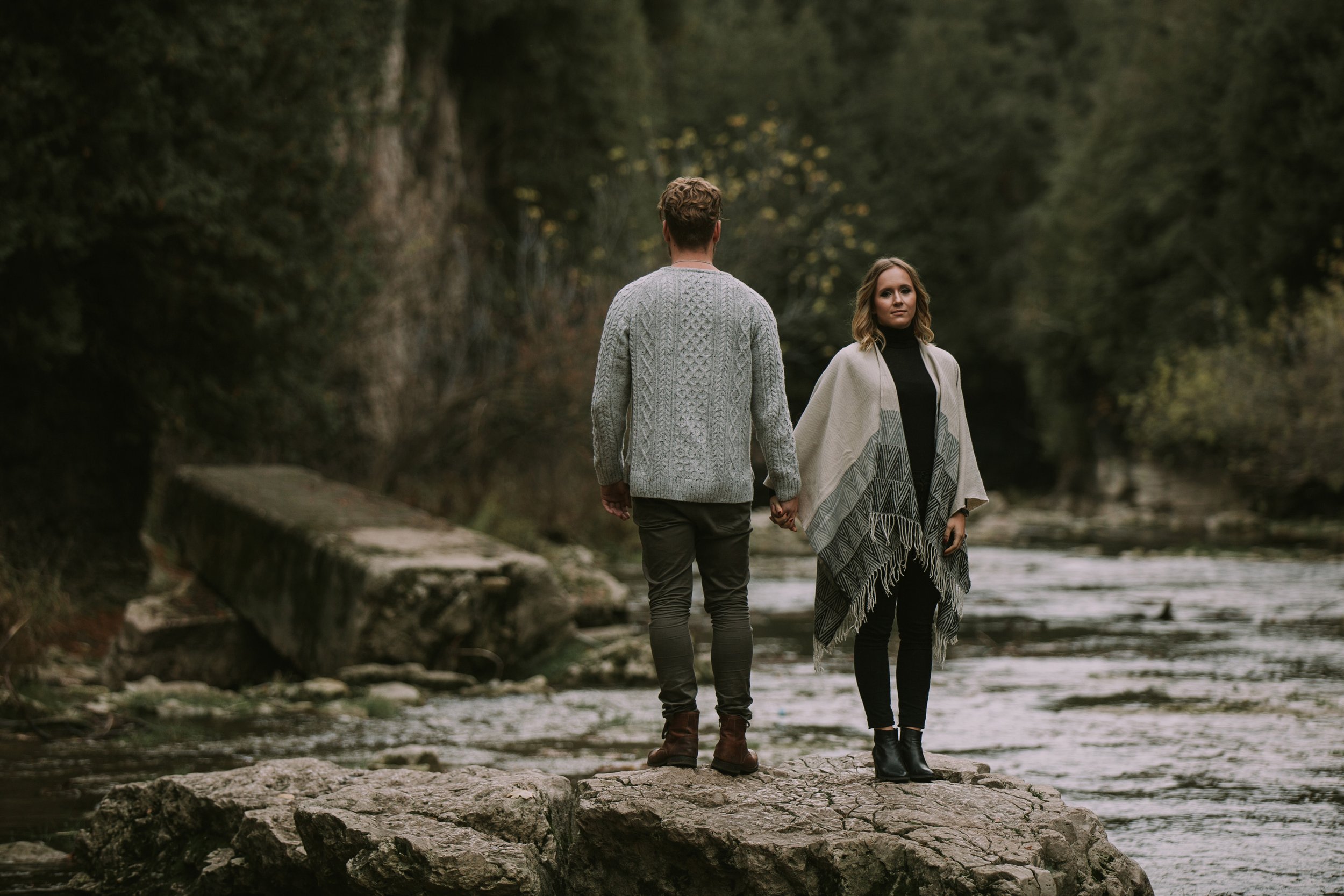 A couple standing back to back on a large rock in a natural autumn setting, holding hands as they look out towards the lush green surroundings.
