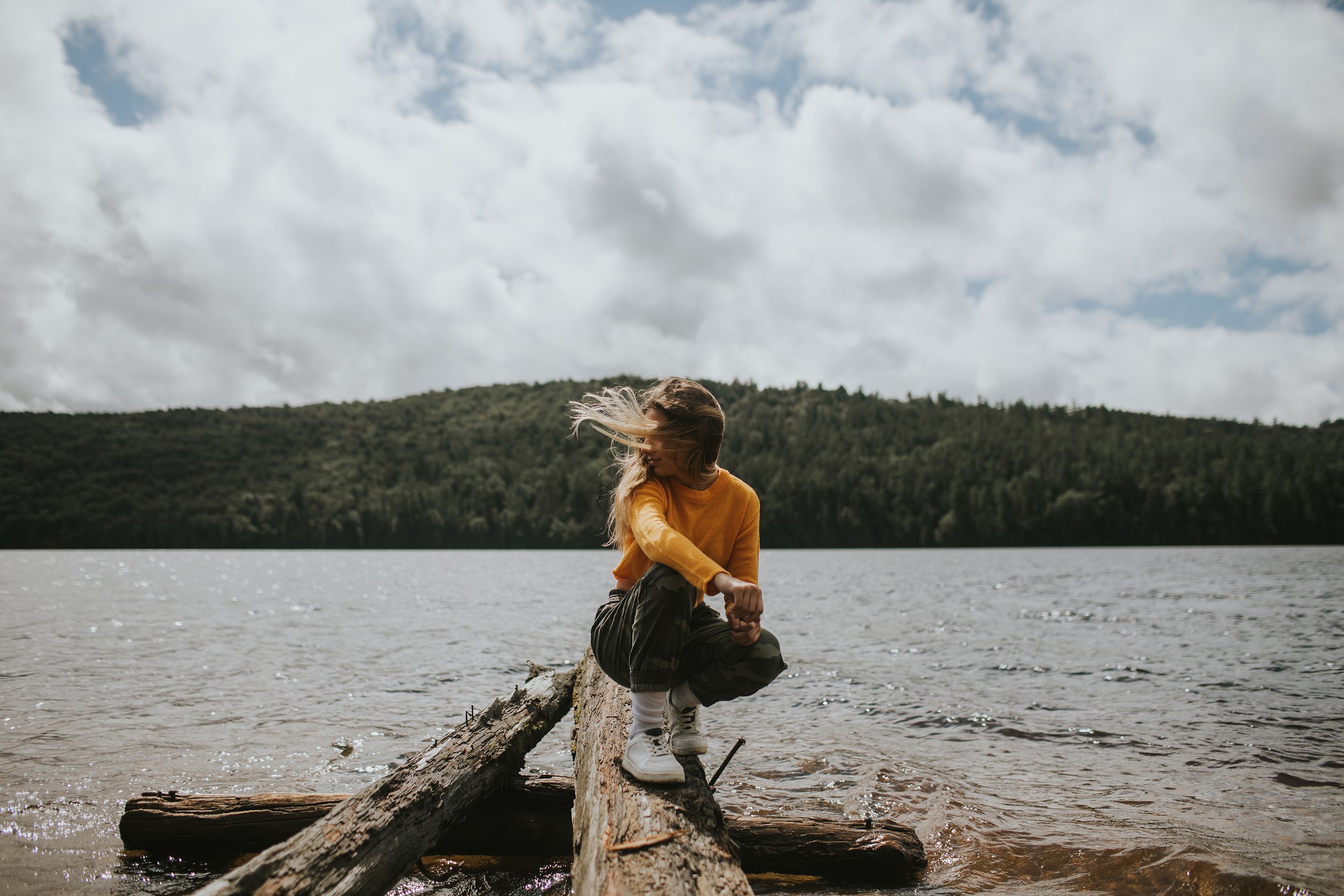 Woman On Log Lake View Nature Portrait London Ontario: A woman in an orange sweater, sitting on a log by a lake, surrounded by a scenic forest view.