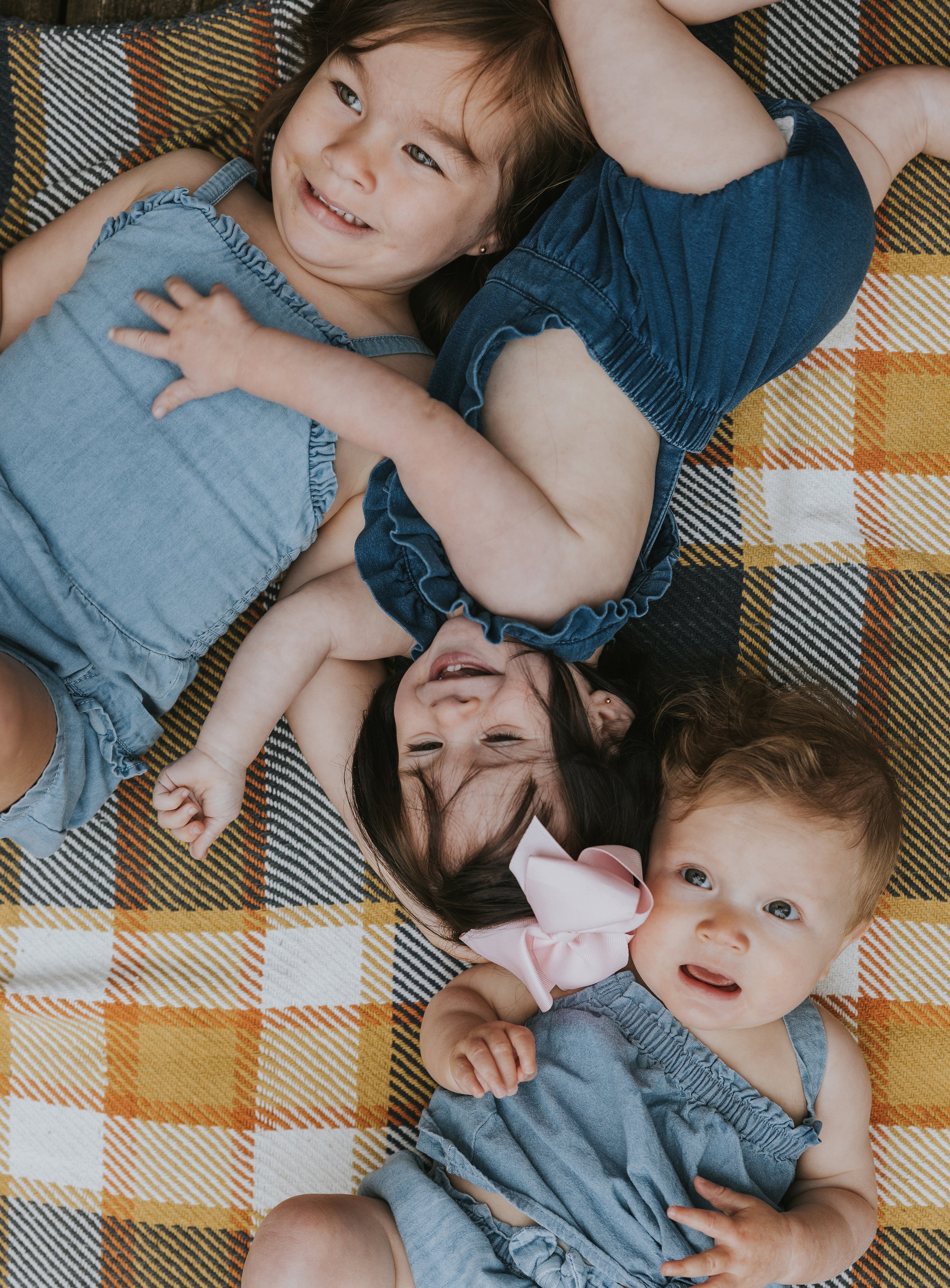 A cheerful young girl lying on a colorful blanket outdoors, smiling widely with two other children in denim outfits.