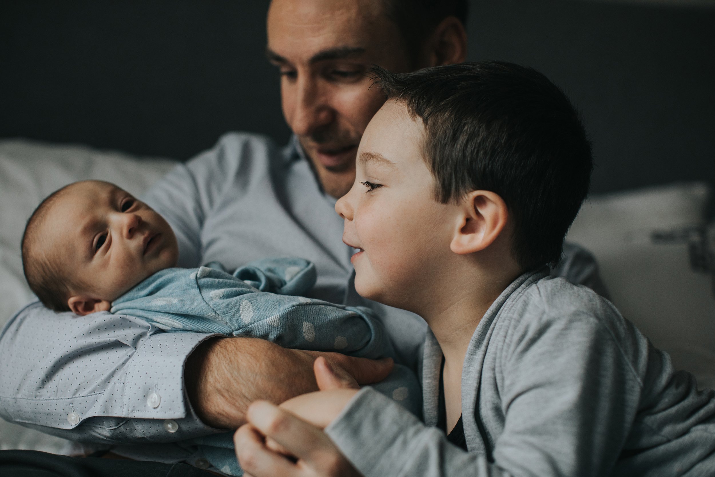 A tender close-up moment of a father holding his newborn baby while the older brother looks on affectionately.
