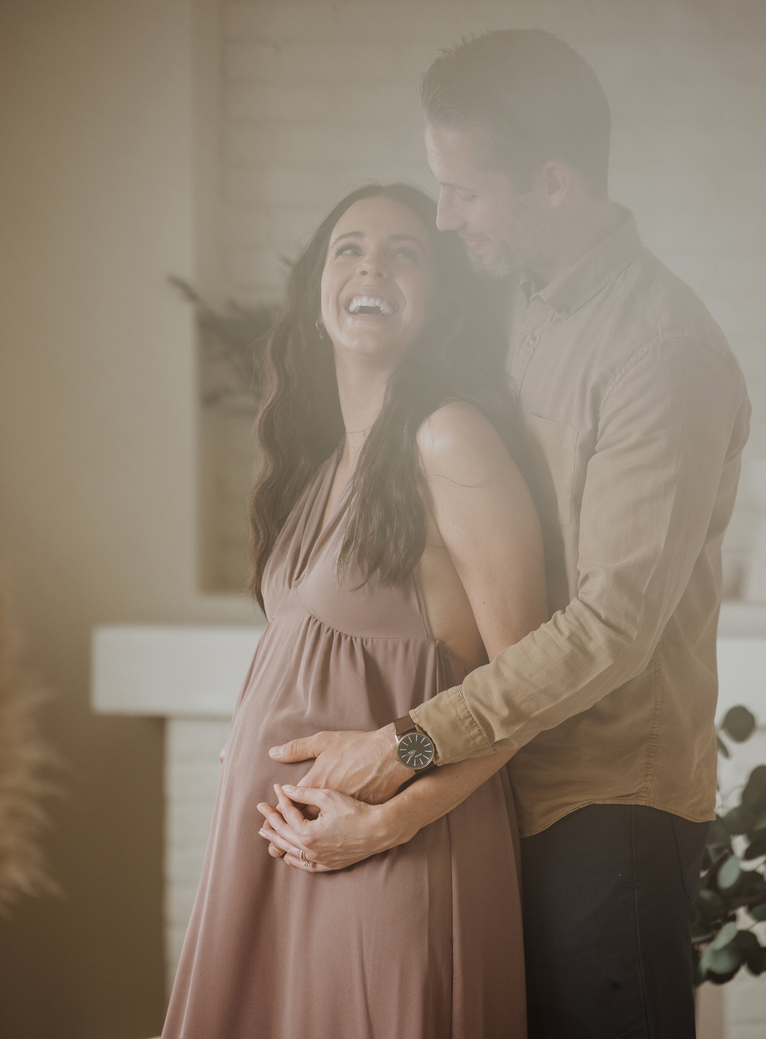 Joyful expectant couple sharing a tender embrace, with the woman laughing and holding her pregnant belly in a soft-lit room.
