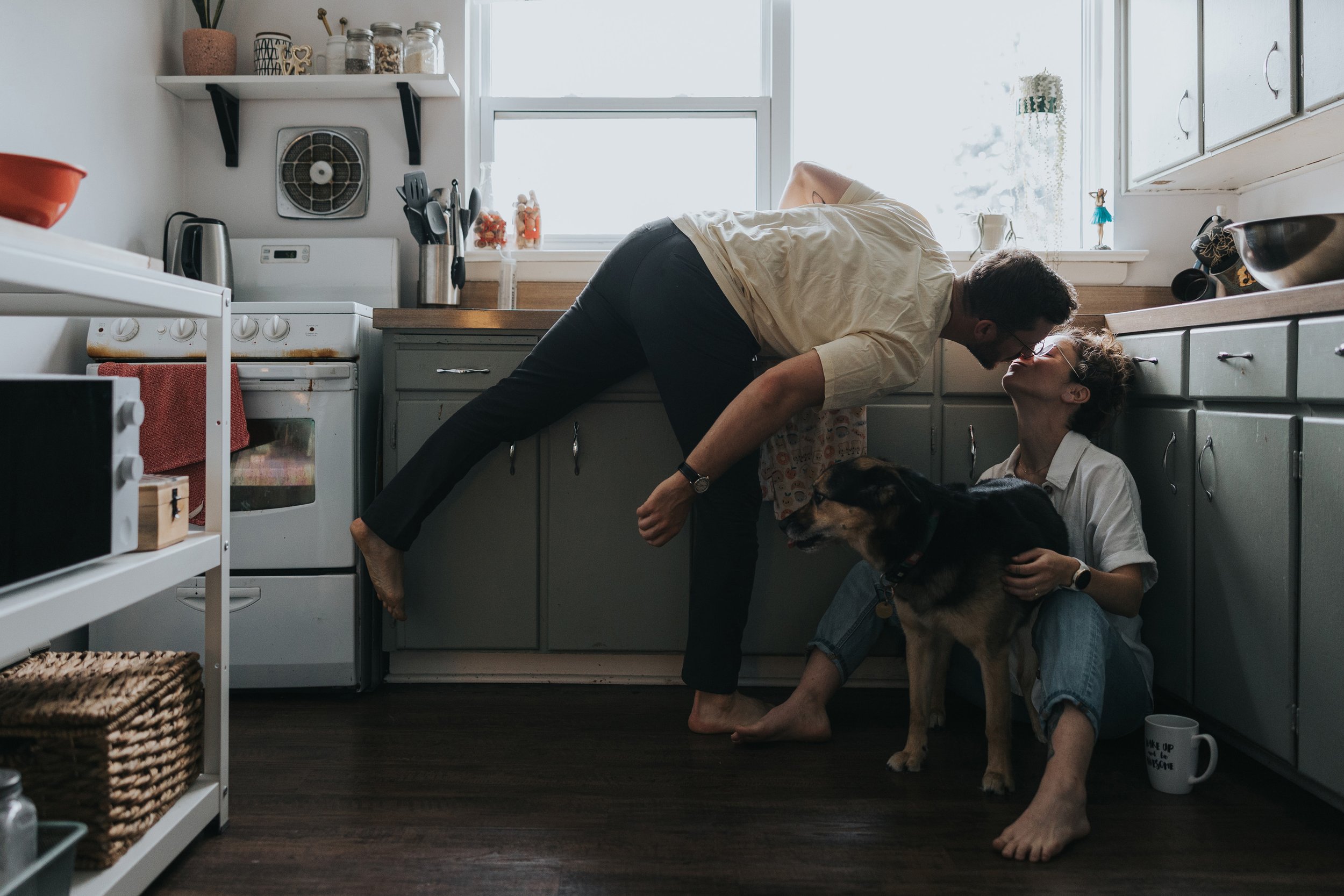 A couple shares a playful moment in the kitchen, with the woman sitting on the floor and the man leaning in for a kiss, while their dog looks on.