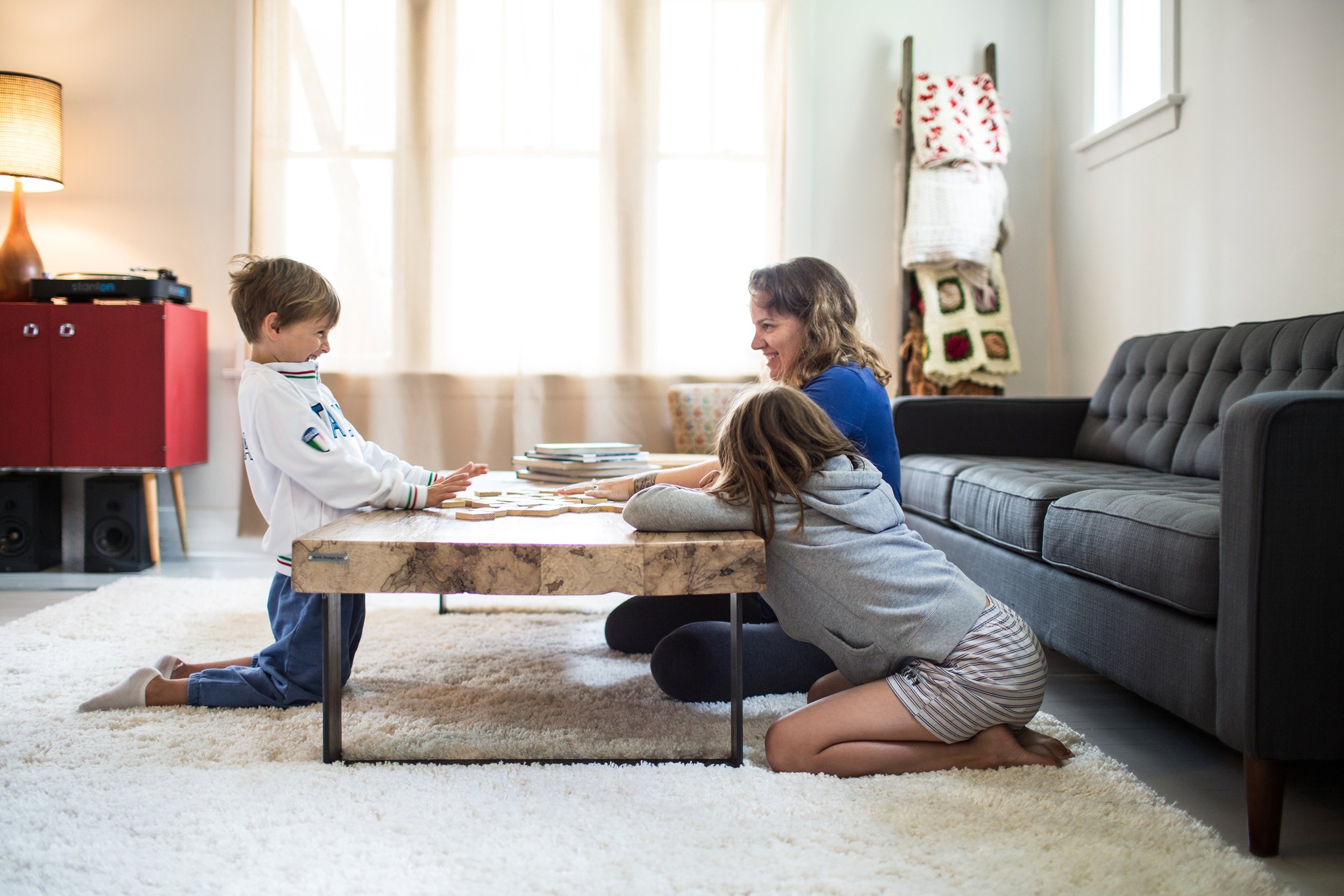 A mother and her two children enjoying a game night at home, gathered around a coffee table in a bright, comfortable living room.