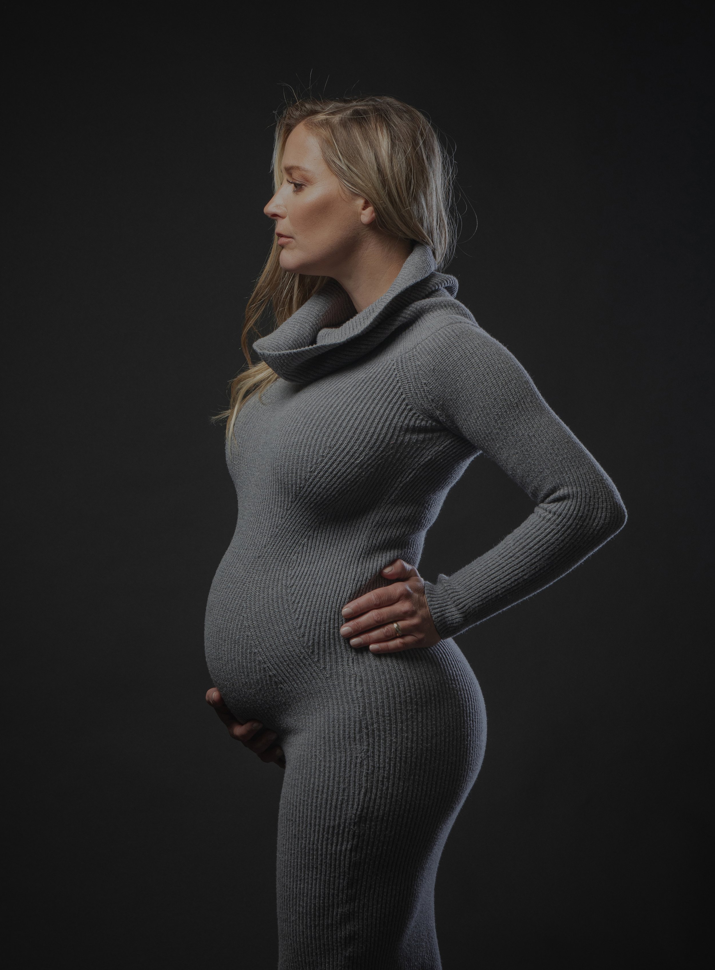 A dramatic studio portrait of a pregnant woman in profile, showcasing her baby bump against a dark background.