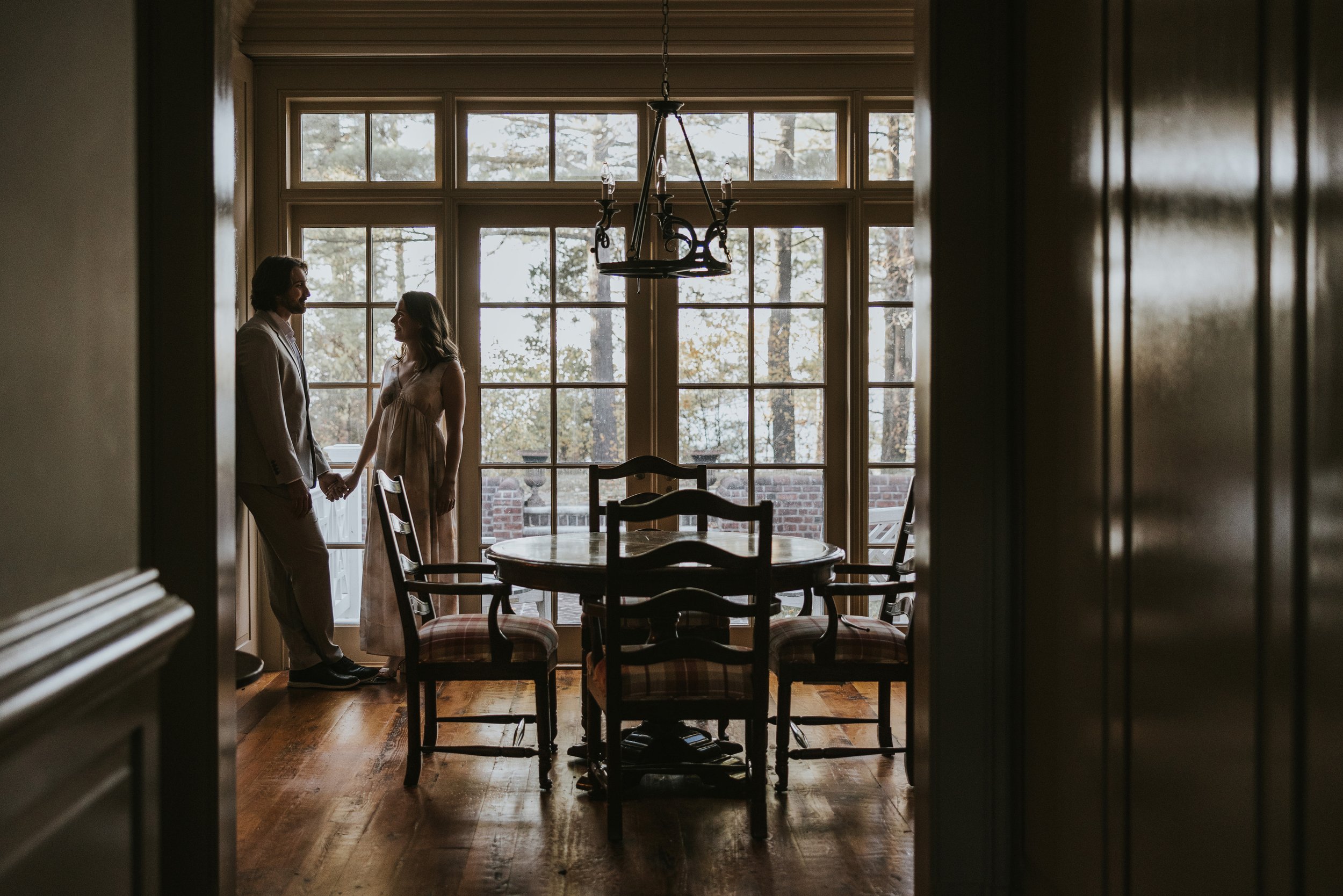 A romantic silhouette of a couple holding hands in a warmly lit dining room, framed by large windows looking out onto a forest.