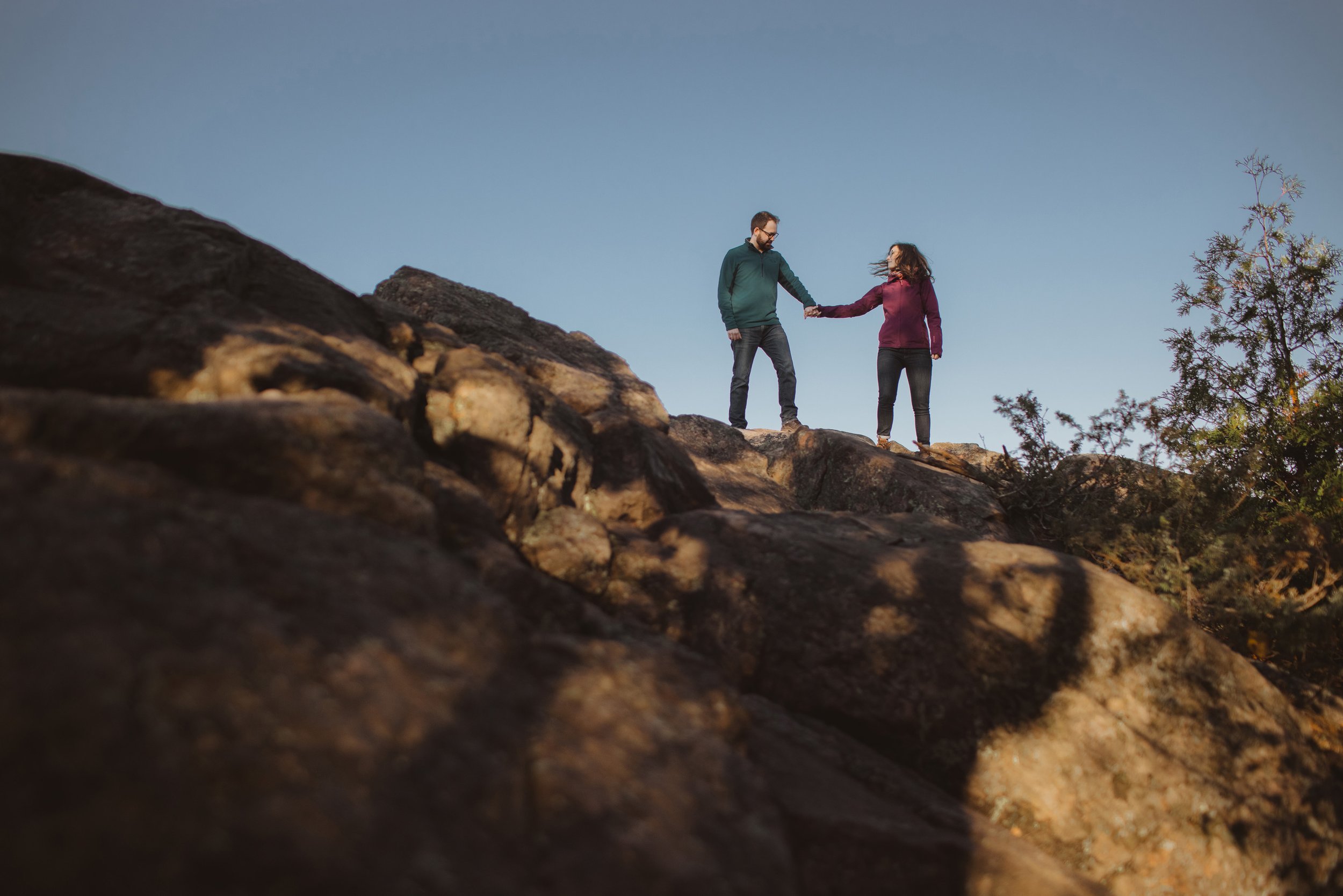 A couple holds hands while standing on a rocky outcrop at Eagle's Nest, bathed in natural light with a clear blue sky above.