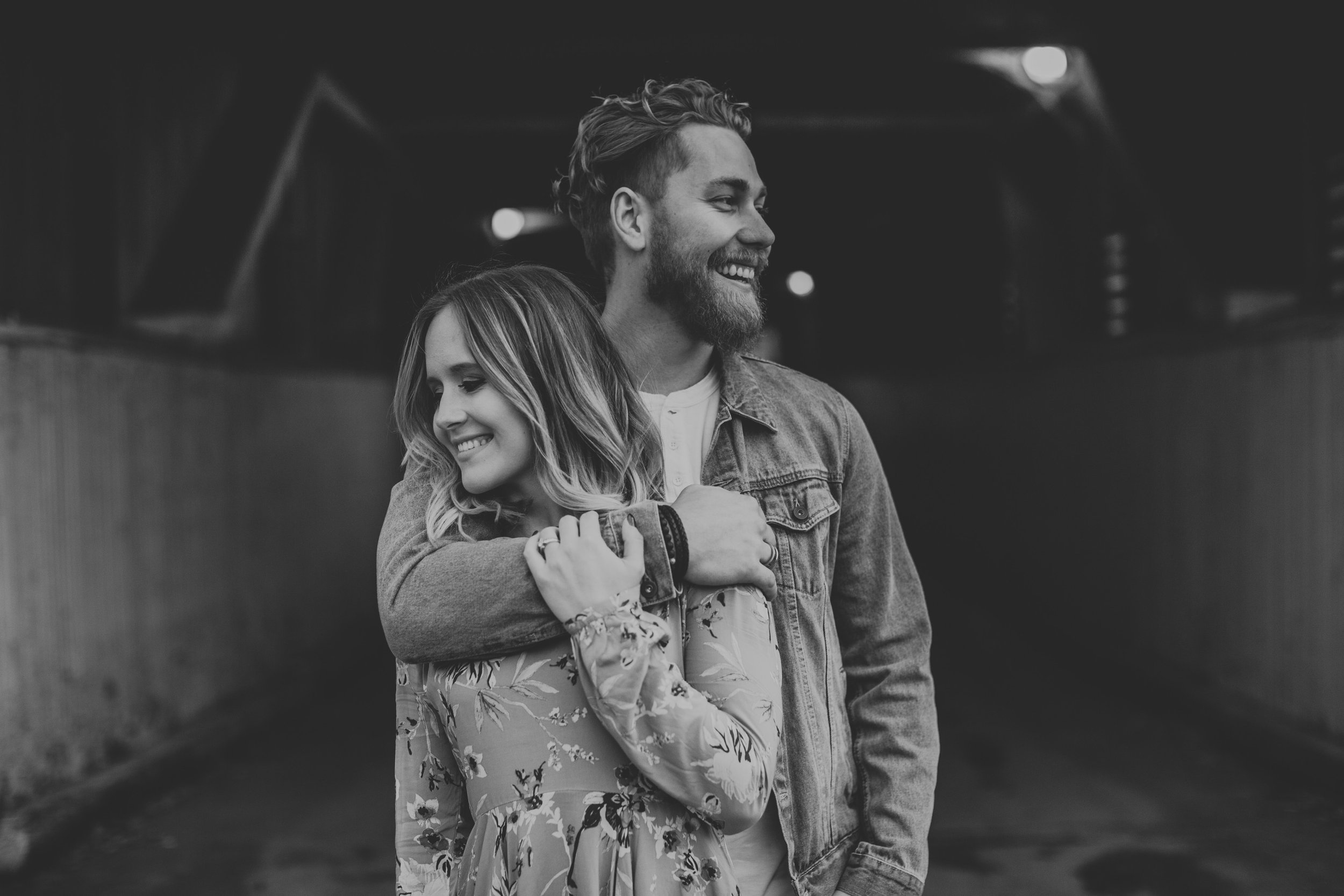 A black-and-white photograph of a couple sharing a joyful embrace in a tunnel during their engagement session. The image conveys a sense of happiness and timeless love.