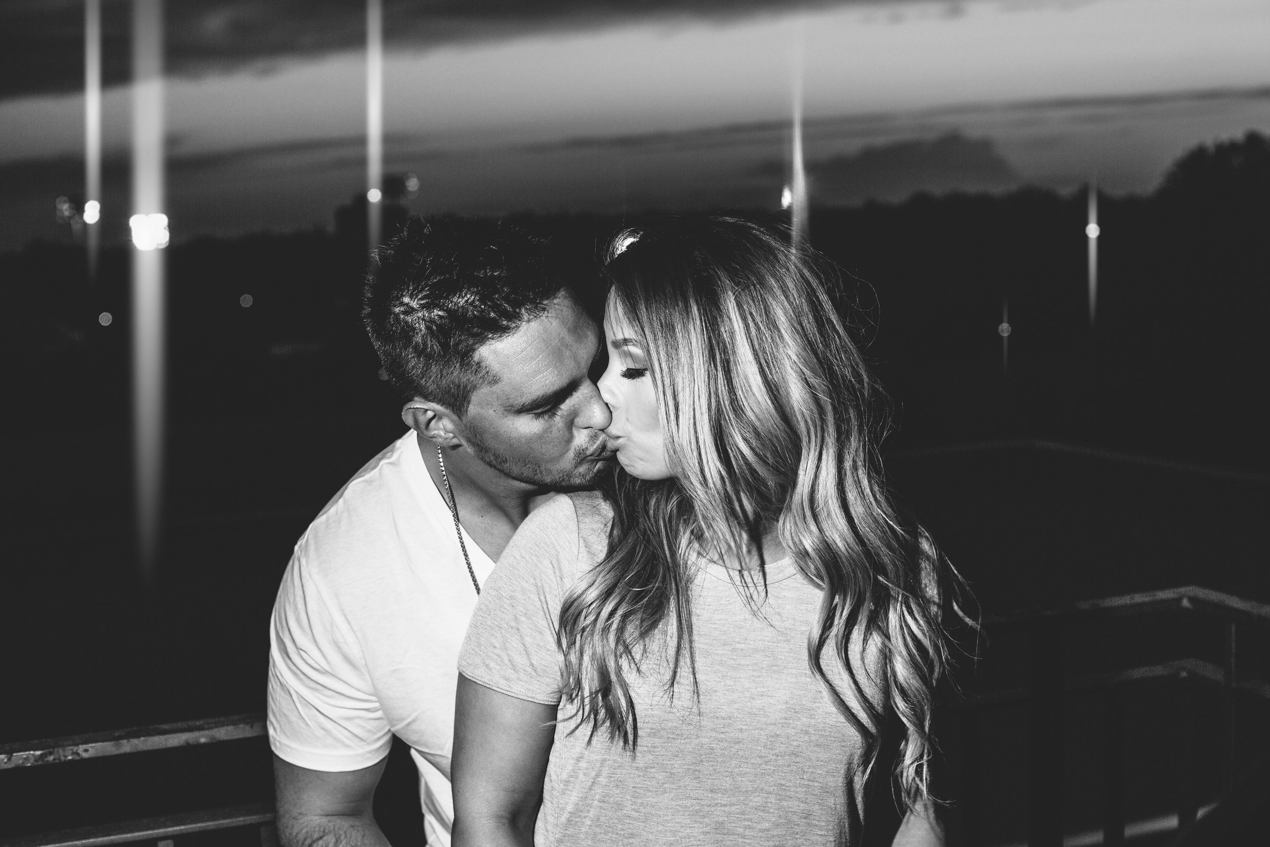 A romantic black-and-white portrait of a couple sharing a kiss during their engagement session at night, illuminated by subtle lights in the background.