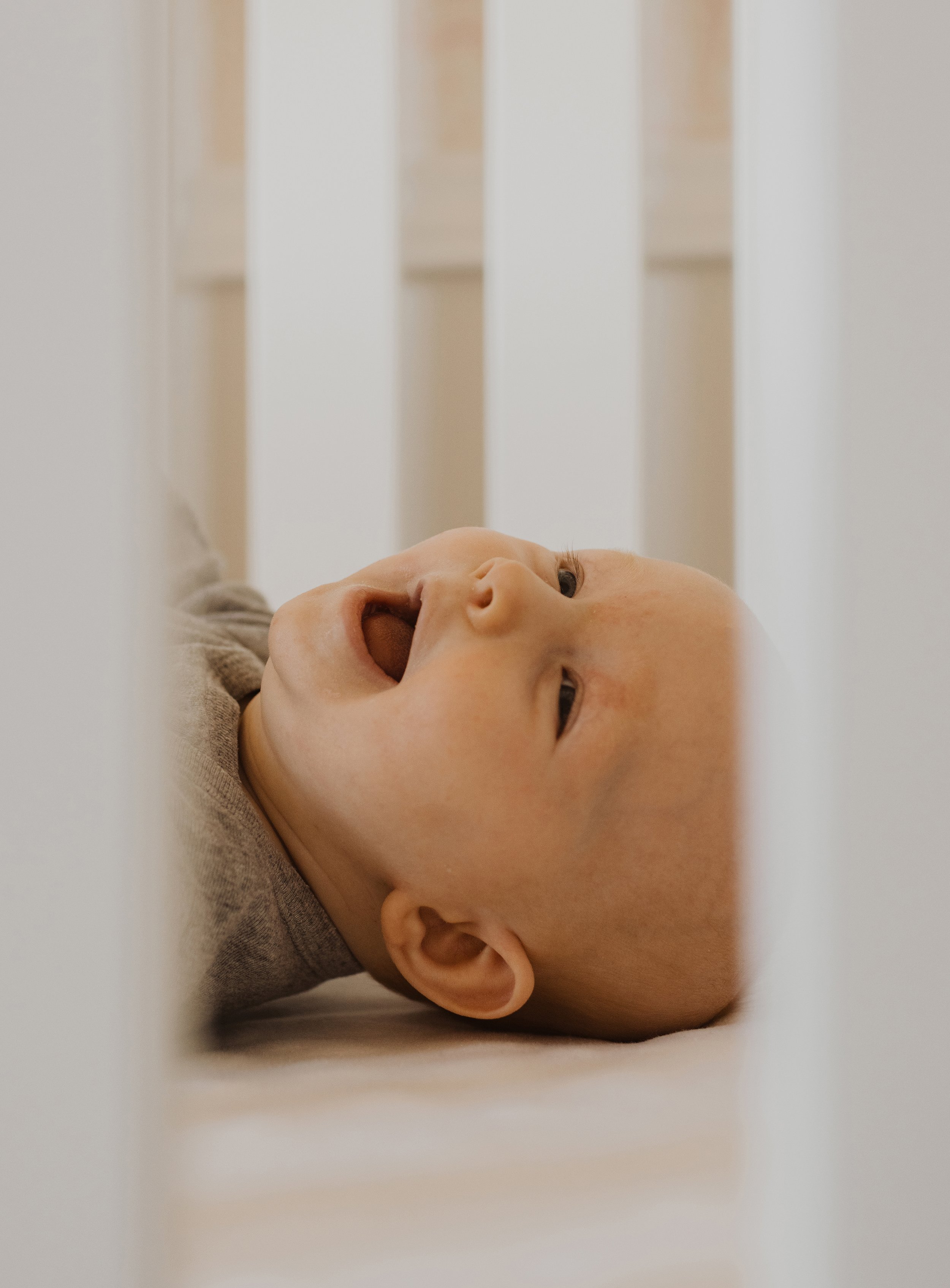 A close-up of a smiling baby lying in a crib, captured from between the bars, with soft lighting enhancing the gentle, joyful expression.