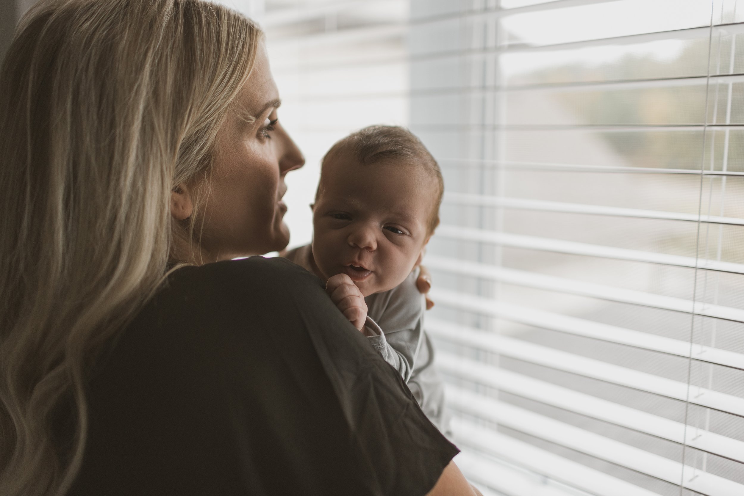 A mother stands by a window holding her newborn, both bathed in soft natural light.