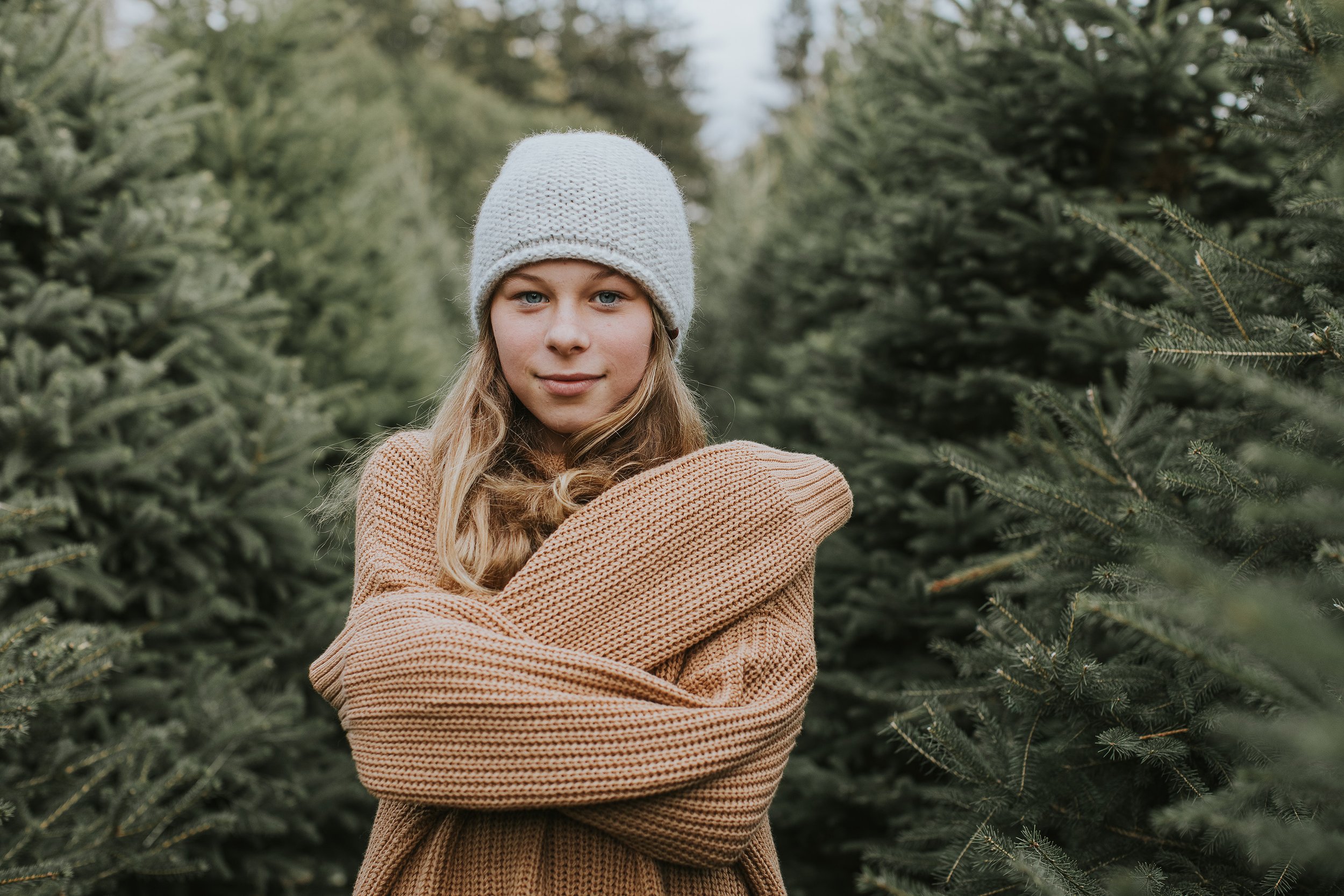 A girl dressed in a cozy sweater and winter hat, standing with her arms crossed in a serene forest of evergreen trees.