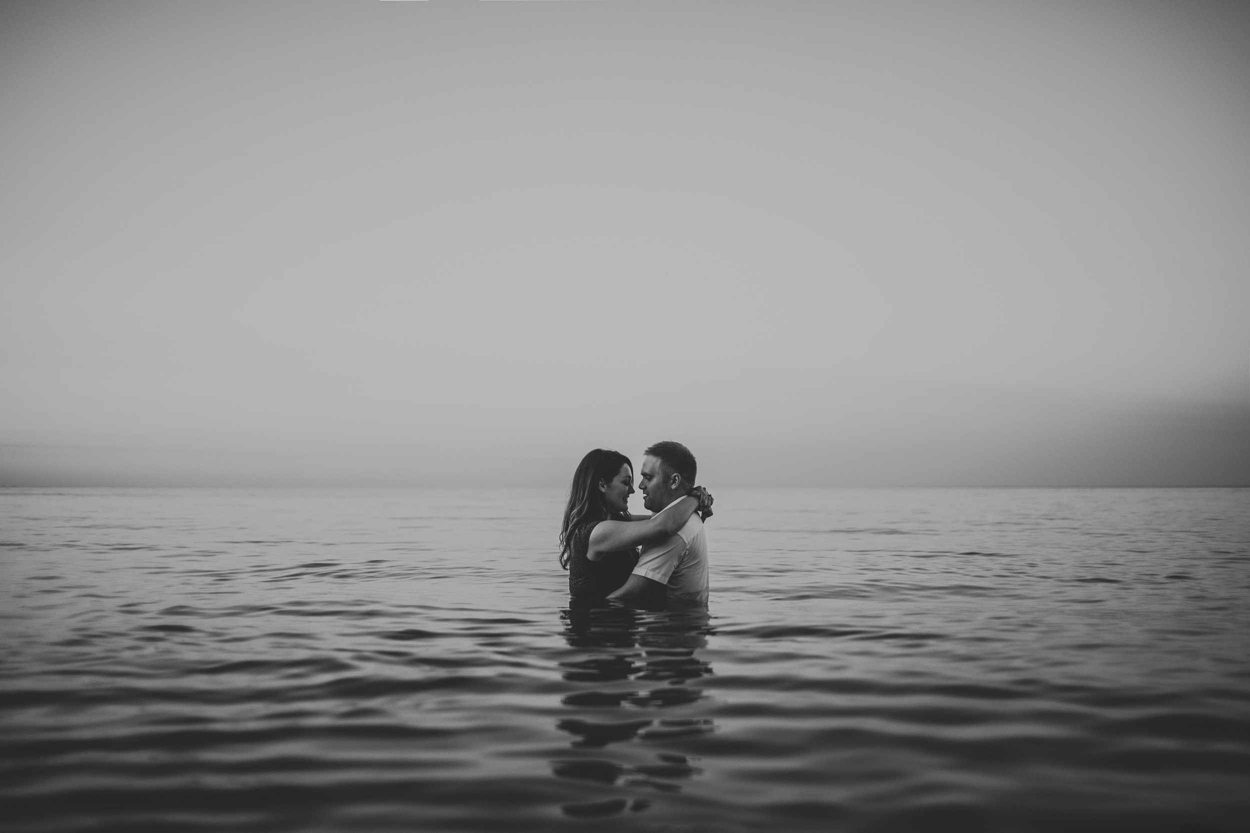 A black and white photo of a couple embracing in the water at sunset, with the serene backdrop highlighting the timeless nature of their love.