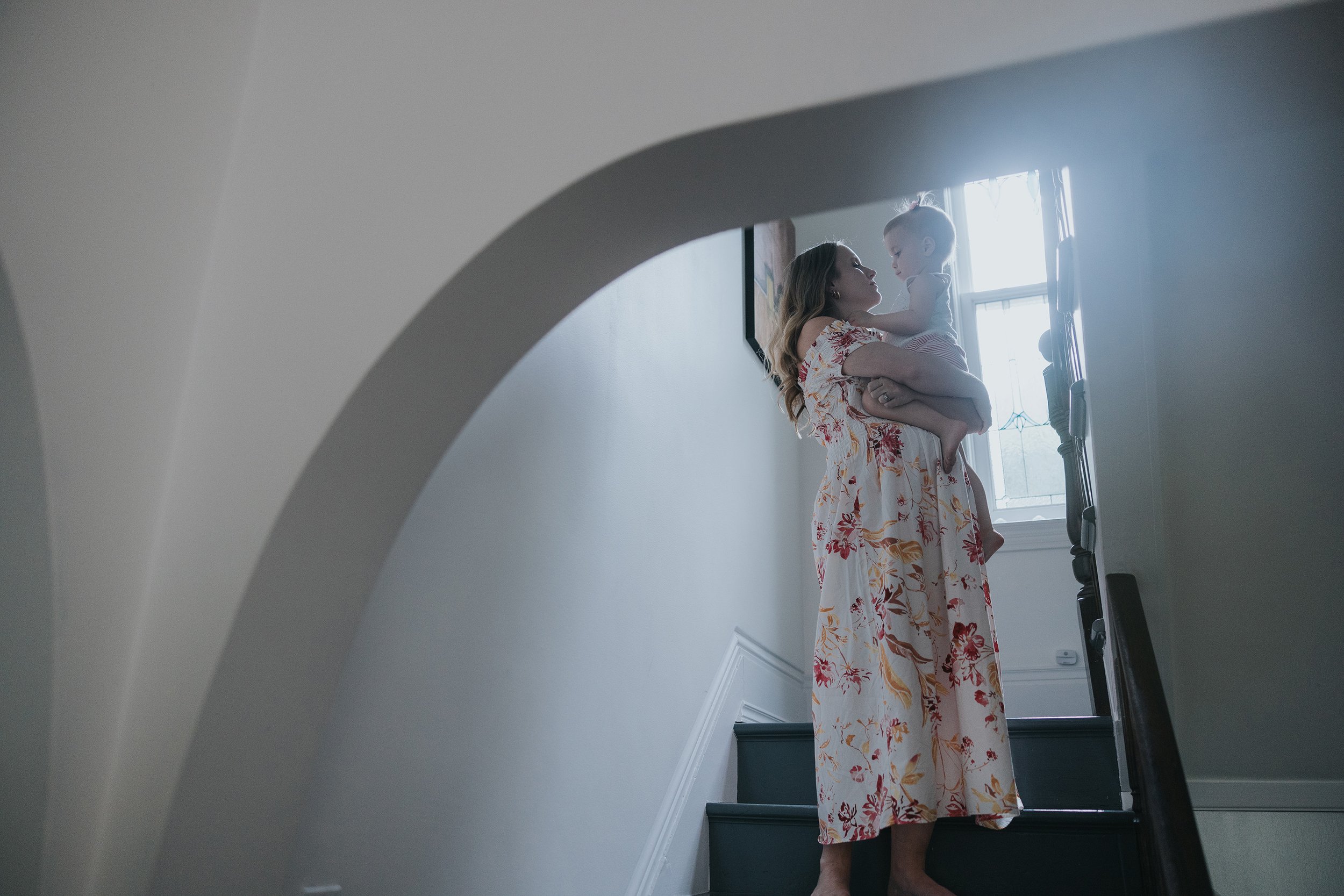 A mother lovingly embraces her baby while standing on a staircase, framed by a soft archway and natural light.