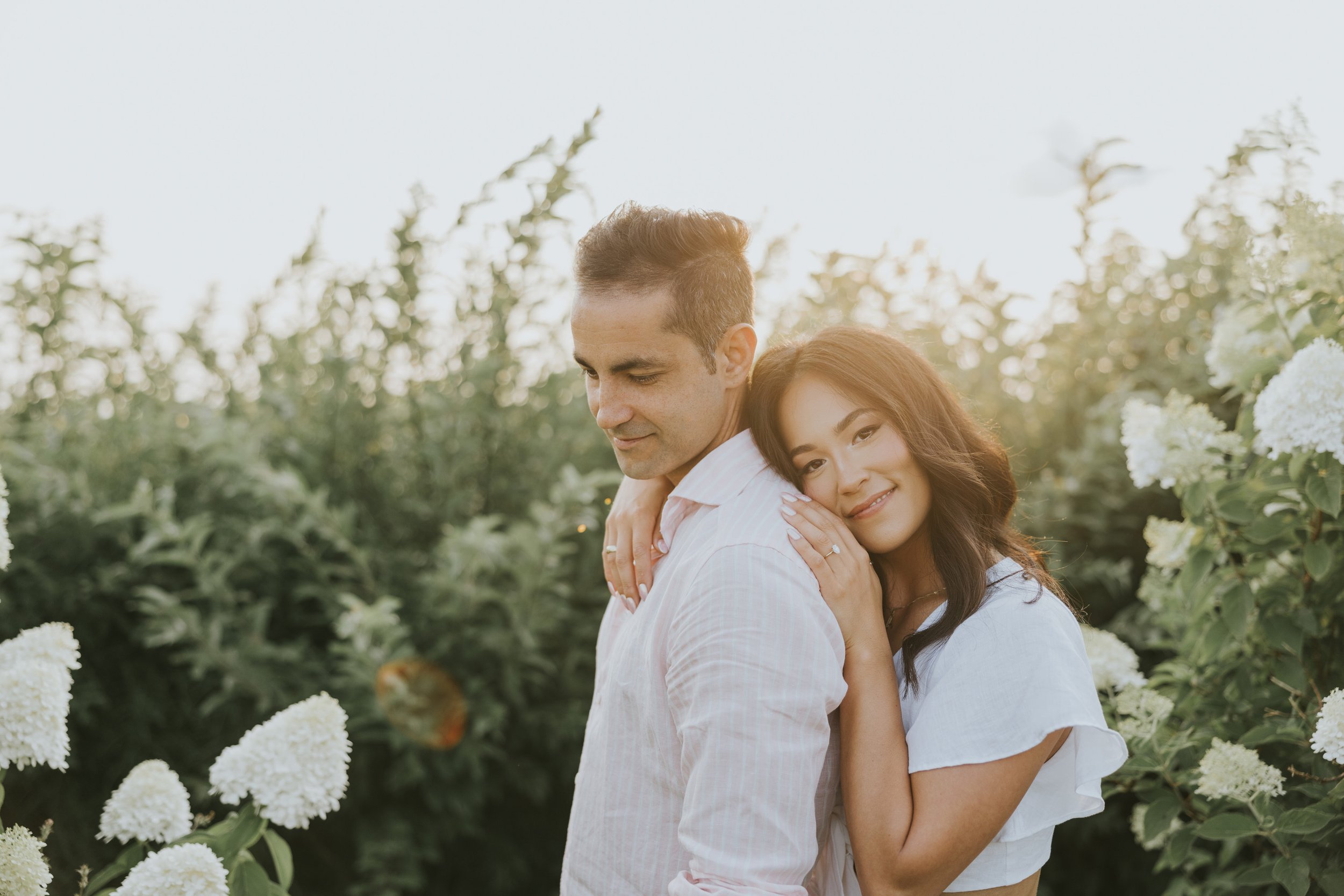 A couple embraces lovingly in a field during golden hour, with soft sunlight illuminating their smiles and the surrounding greenery.
