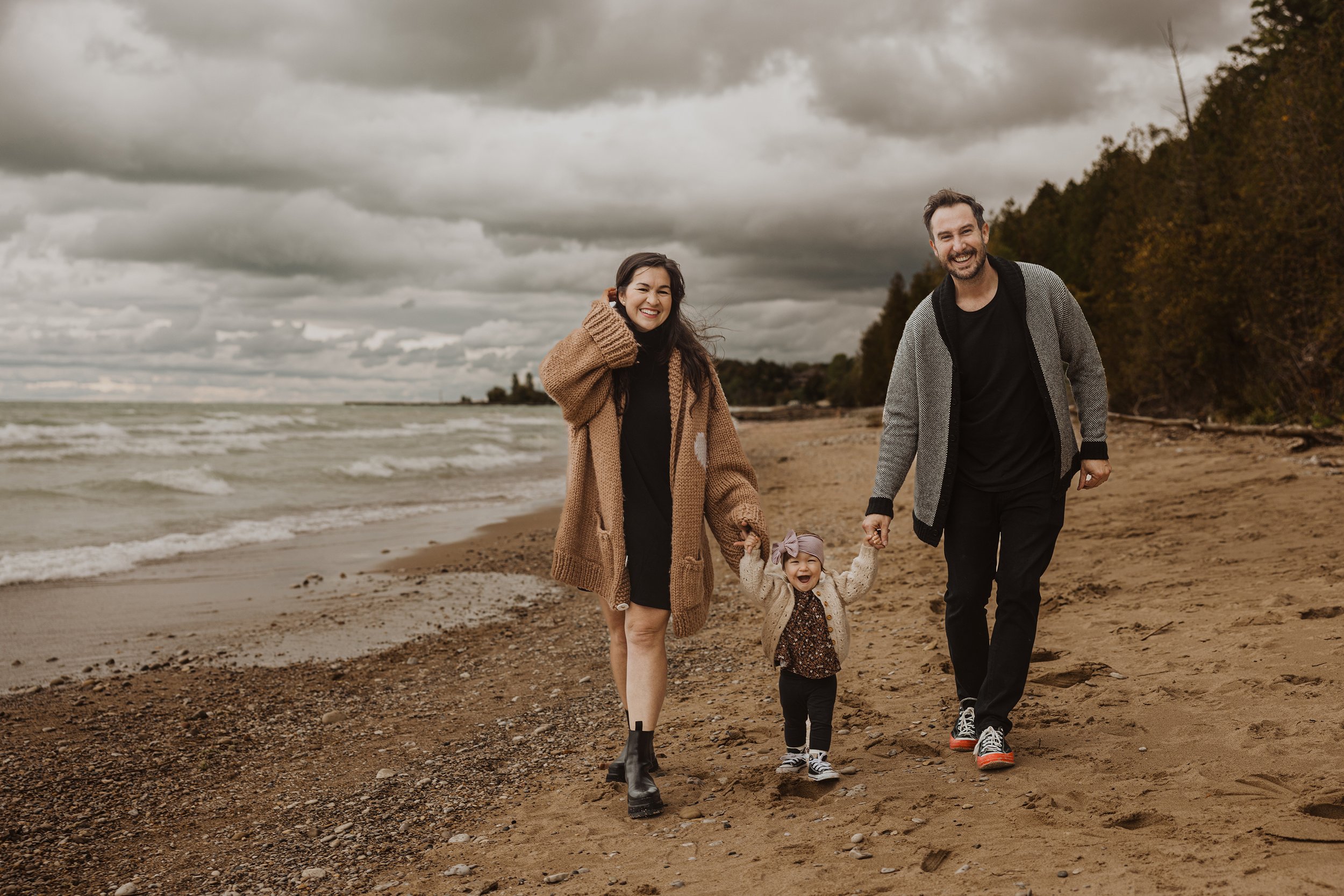 A family of three walking along a moody beach under stormy skies, holding hands and smiling as their daughter swings between them.