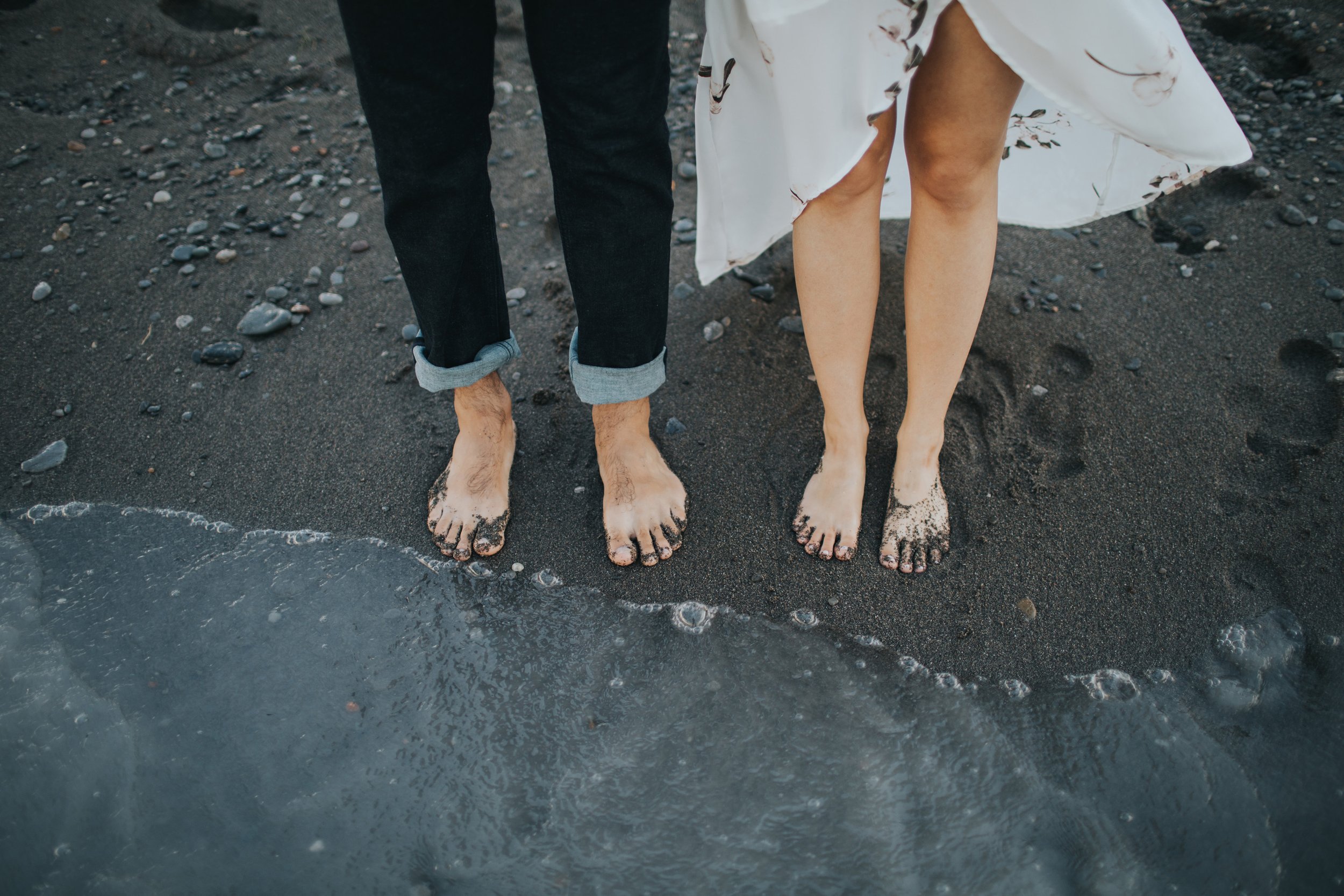 Close-up detail of a couple's barefoot feet standing in the sand at the edge of the water during a beach engagement session. The gentle waves add a soothing touch to the scene.