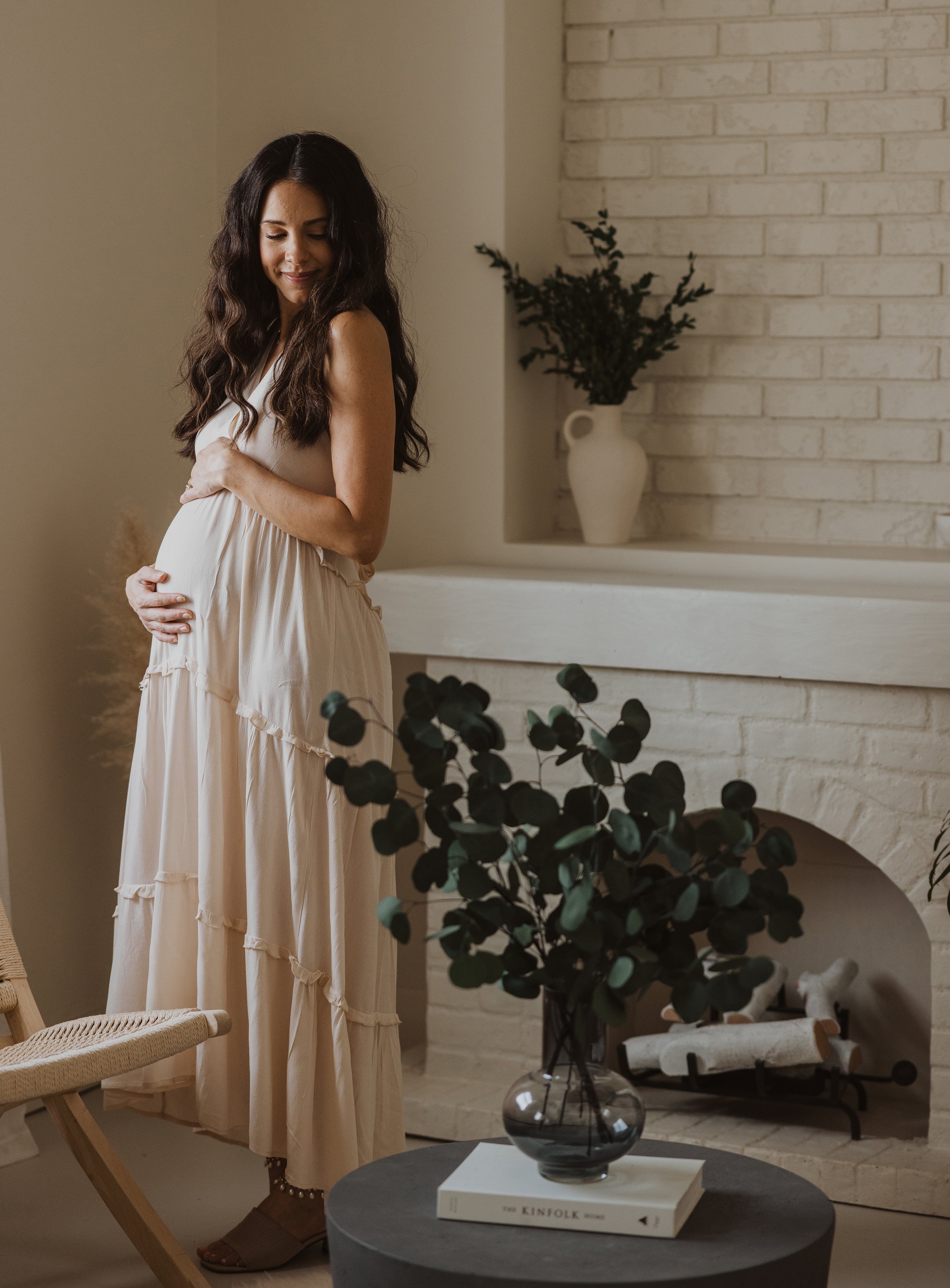 A serene indoor maternity portrait of a woman standing by a vase of greenery, gently holding her baby bump.