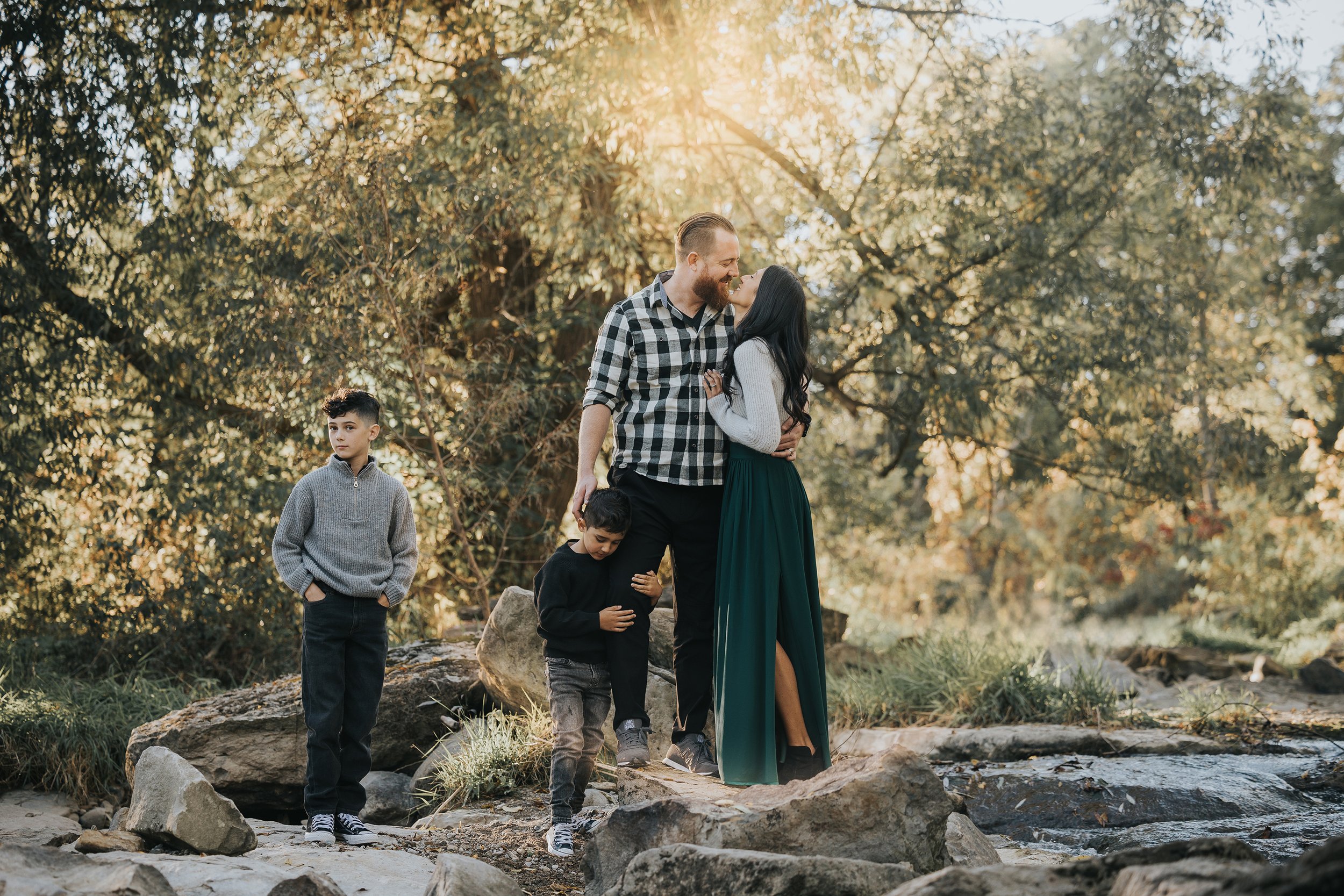 A romantic family portrait in nature, where the parents share a kiss while their two young sons explore the surroundings.