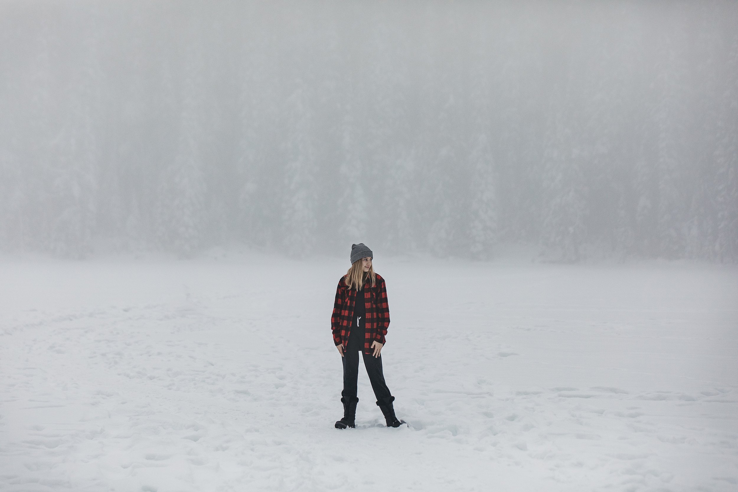 A lone figure standing in a snow-covered, foggy wilderness, wearing a red and black checkered jacket, with trees faintly visible in the background.