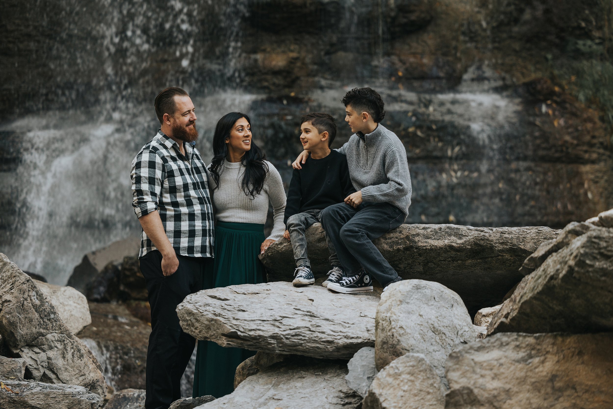This is a family portrait taken near a waterfall, with the family standing or sitting on large rocks. The parents and two young boys are all dressed in cozy, casual clothing, and the boys are engaging with their parents, creating a candid, joyful mom