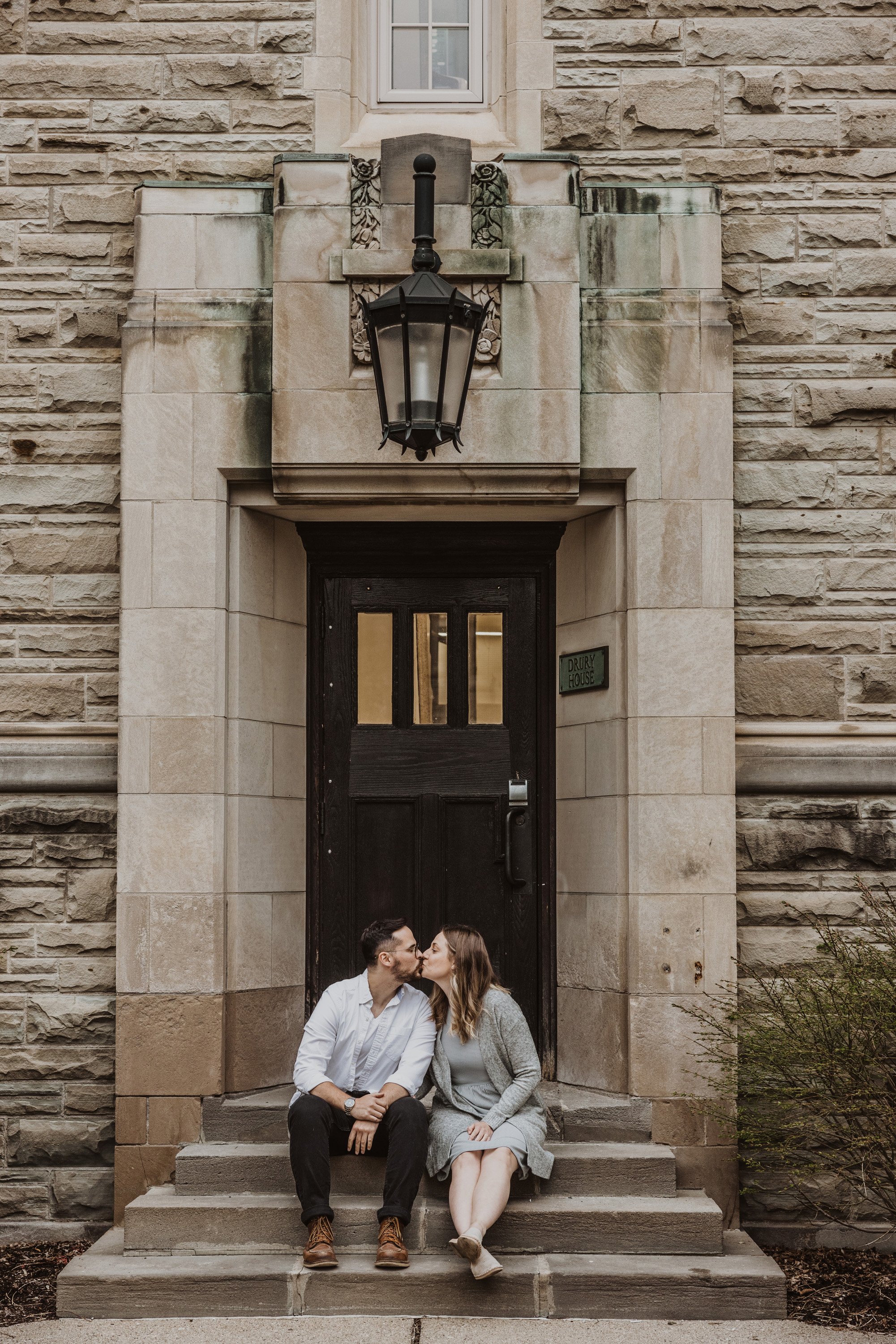 A couple kisses while sitting on stone entrance stairs, their closeness highlighted by the historic architectural backdrop.