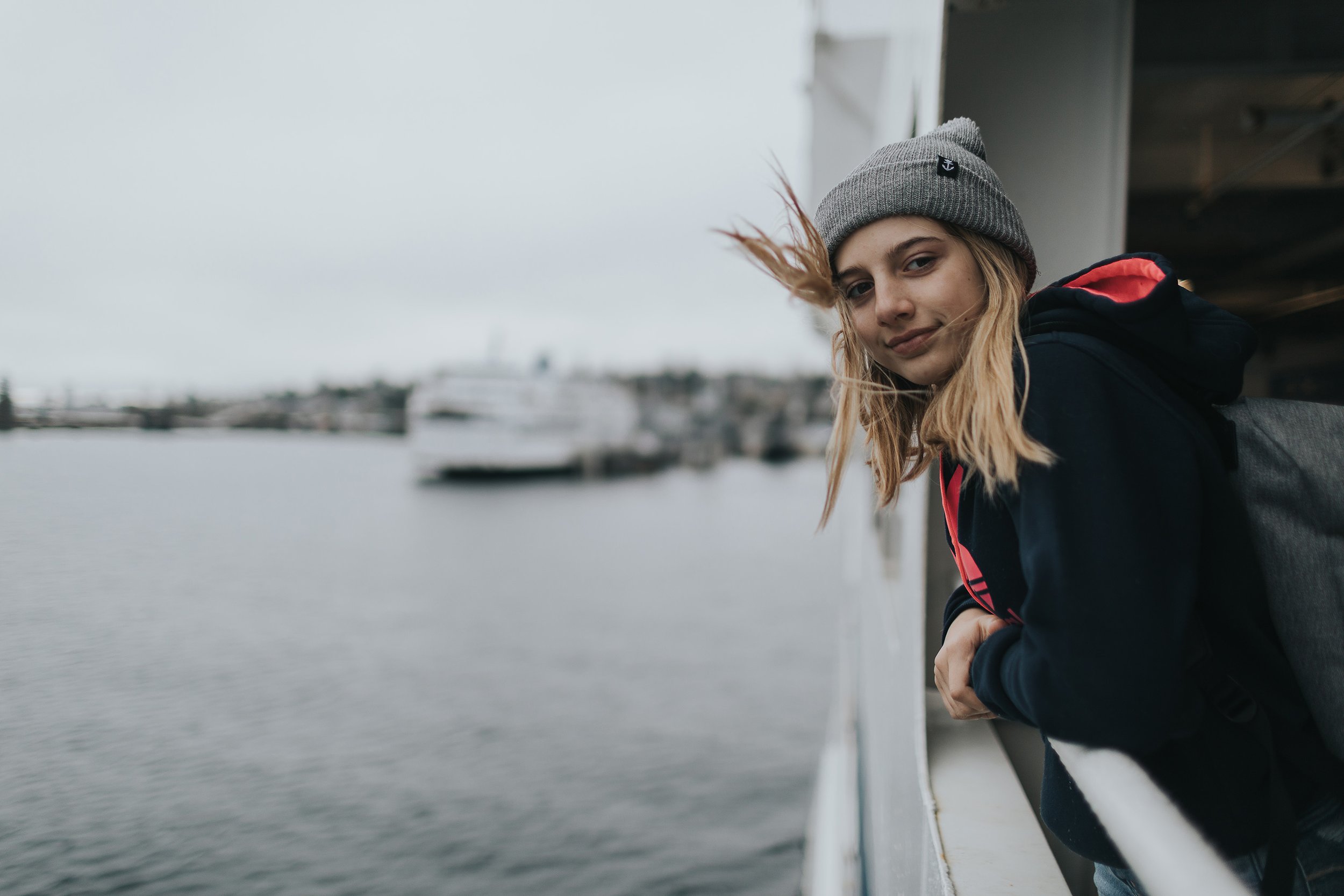 A teenager stands on the deck of a ferry boat, with the wind blowing through their hair, looking out over a harbour.
