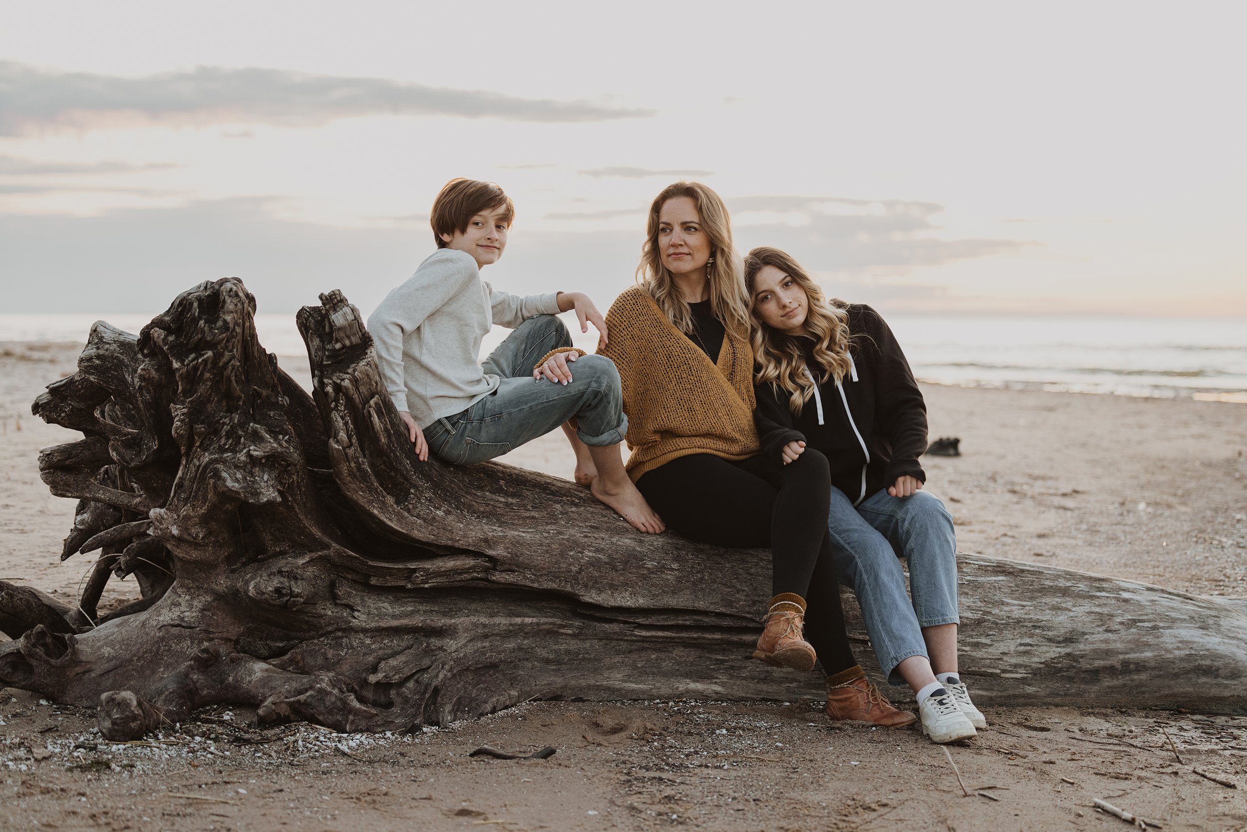 A casual portrait of a mother with her two children, sitting on a large piece of driftwood at the beach.