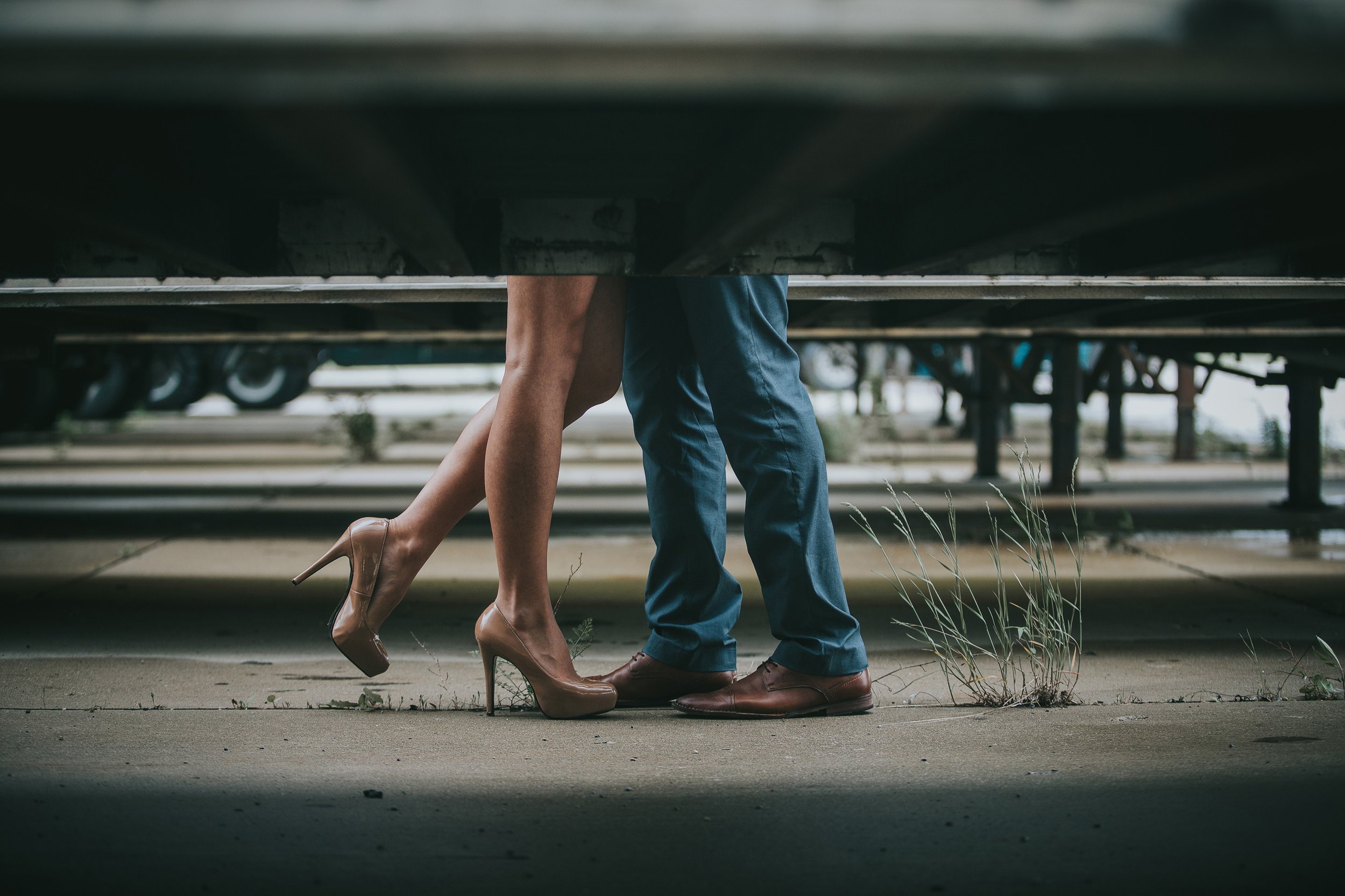 A couple shares a kiss underneath a truck, the industrial urban setting contrasting with their tender moment of connection.