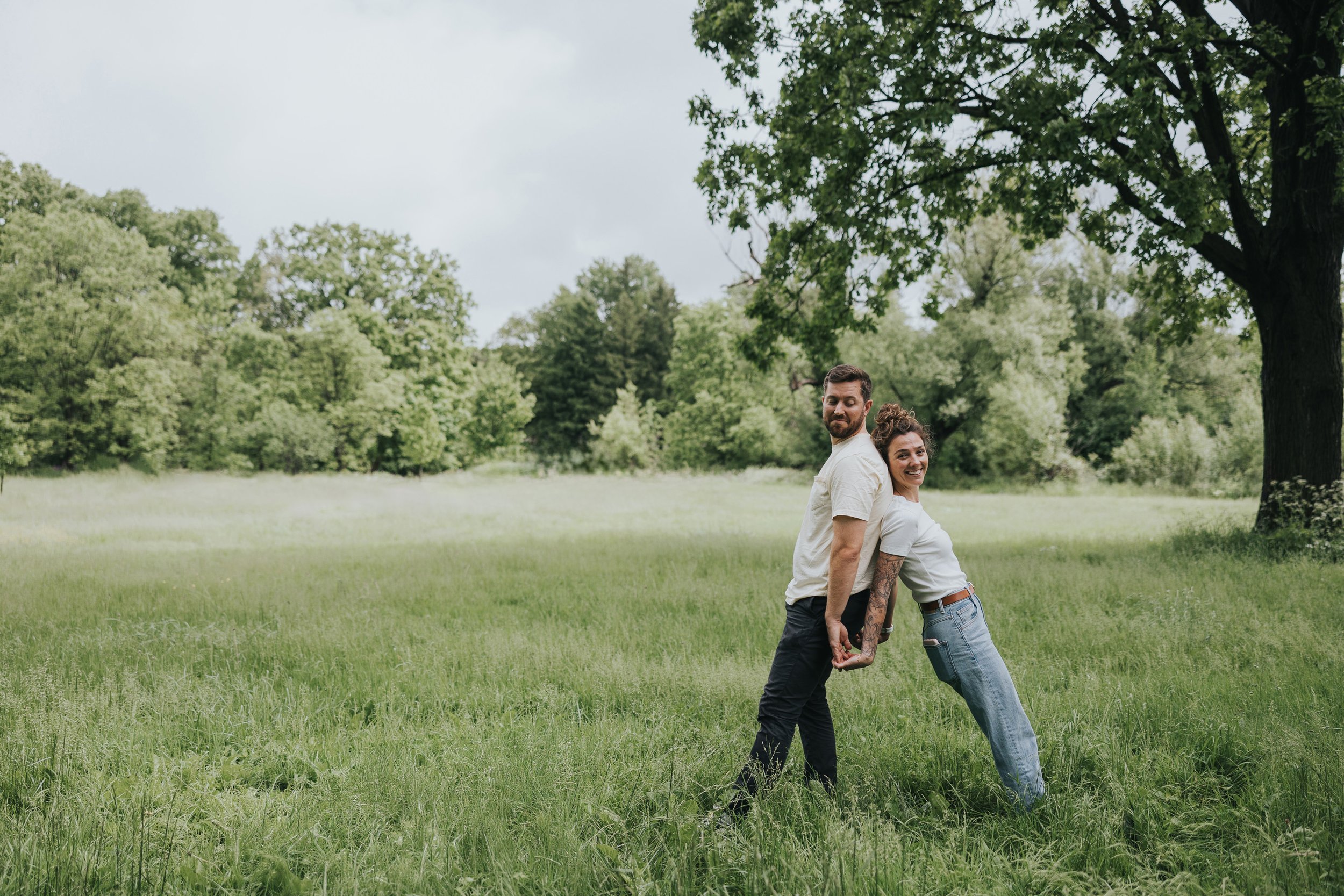 A couple stands back to back in a grassy meadow, holding hands and leaning away from each other in a playful pose.