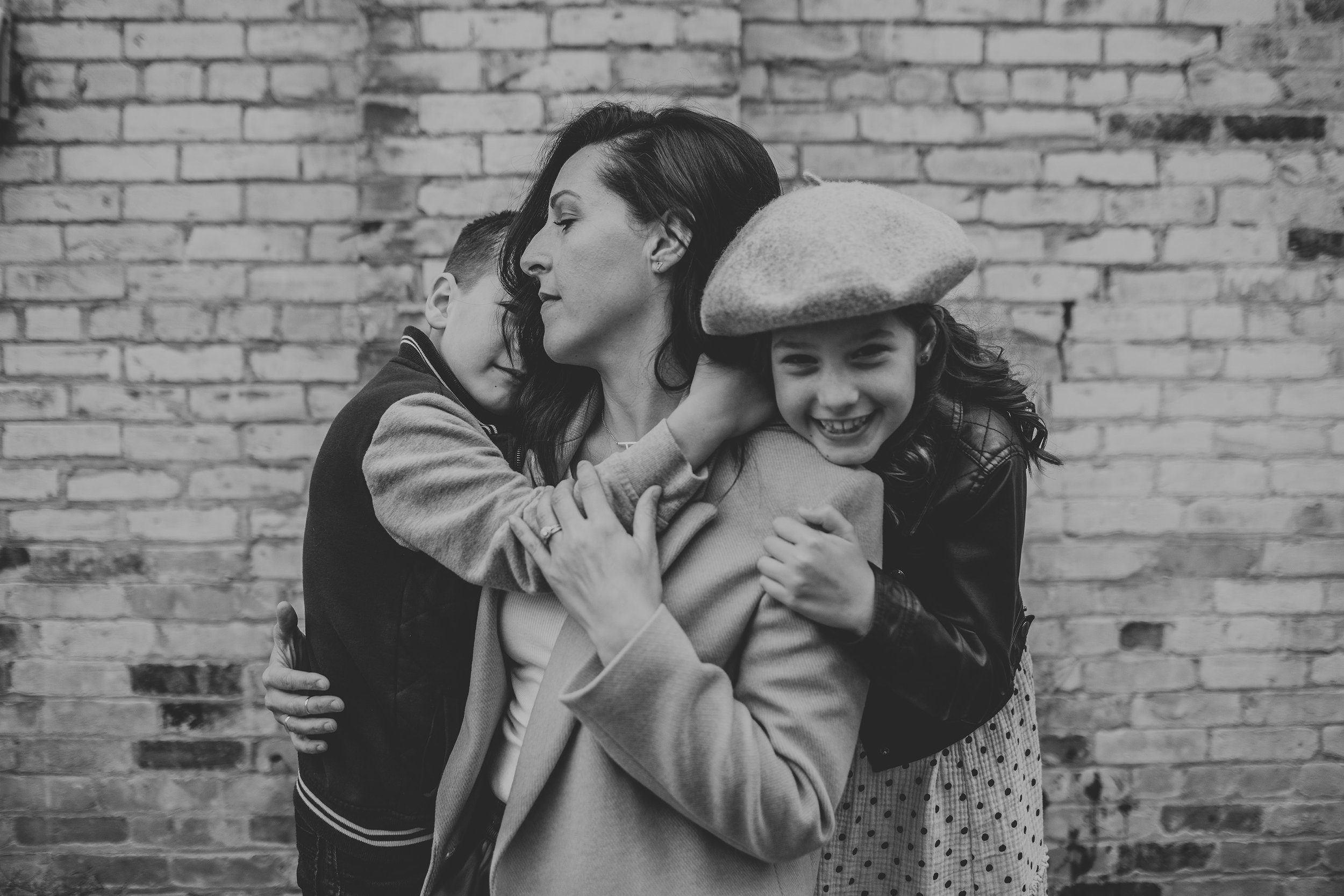 A candid moment of a mother hugging her two children close, standing against an urban brick wall.