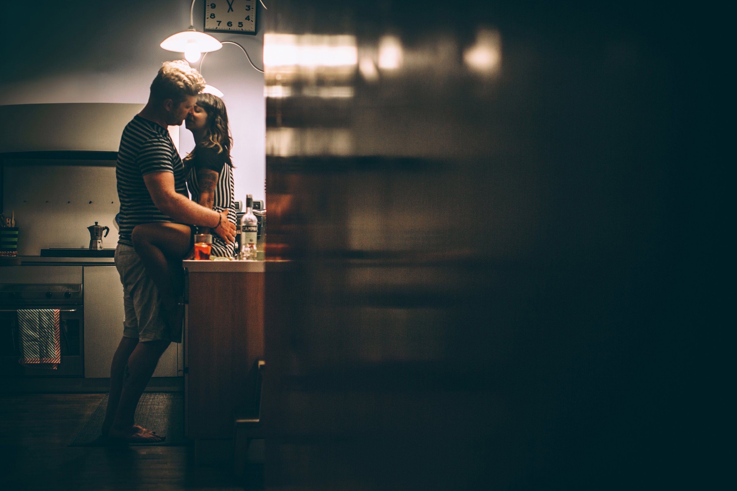 A couple shares an intimate moment in a dimly lit kitchen, the warm lighting accentuating the romantic atmosphere.