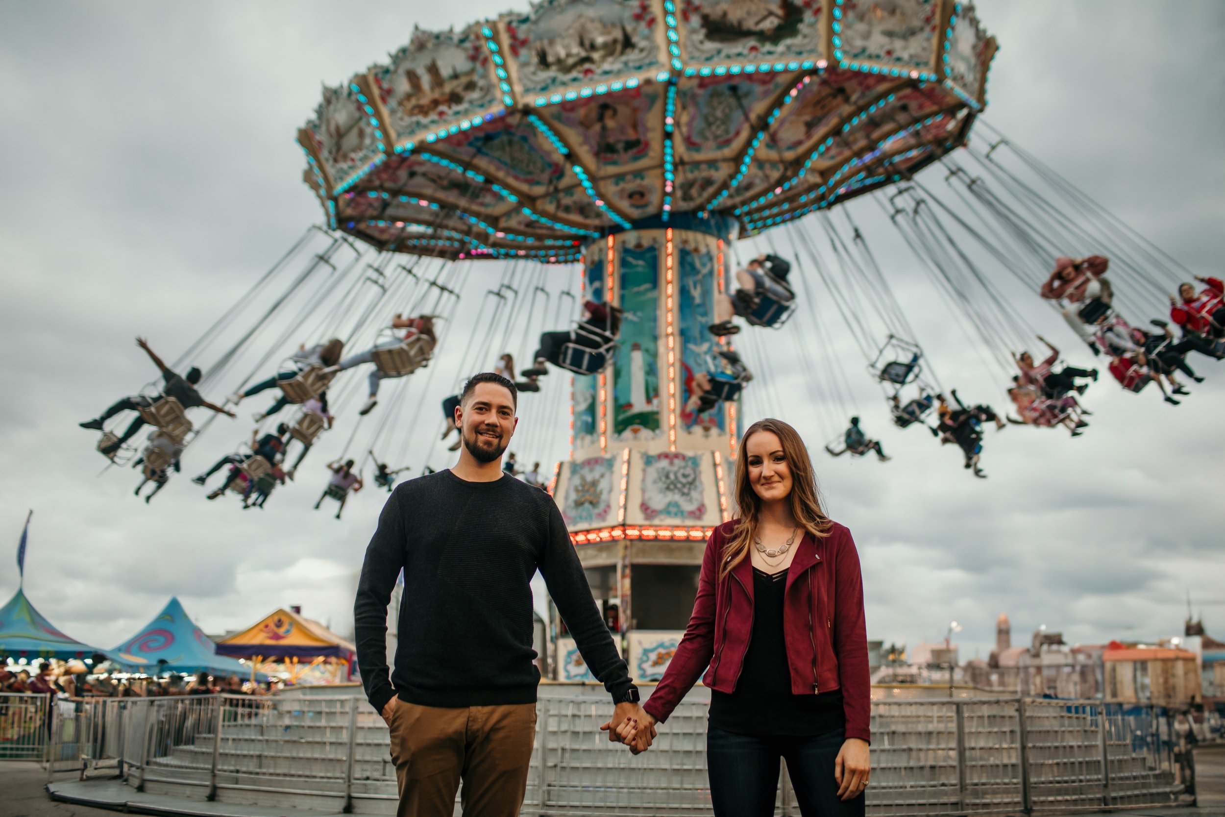 A couple stands hand in hand in front of a brightly lit carnival swing ride, enjoying a festive atmosphere together.