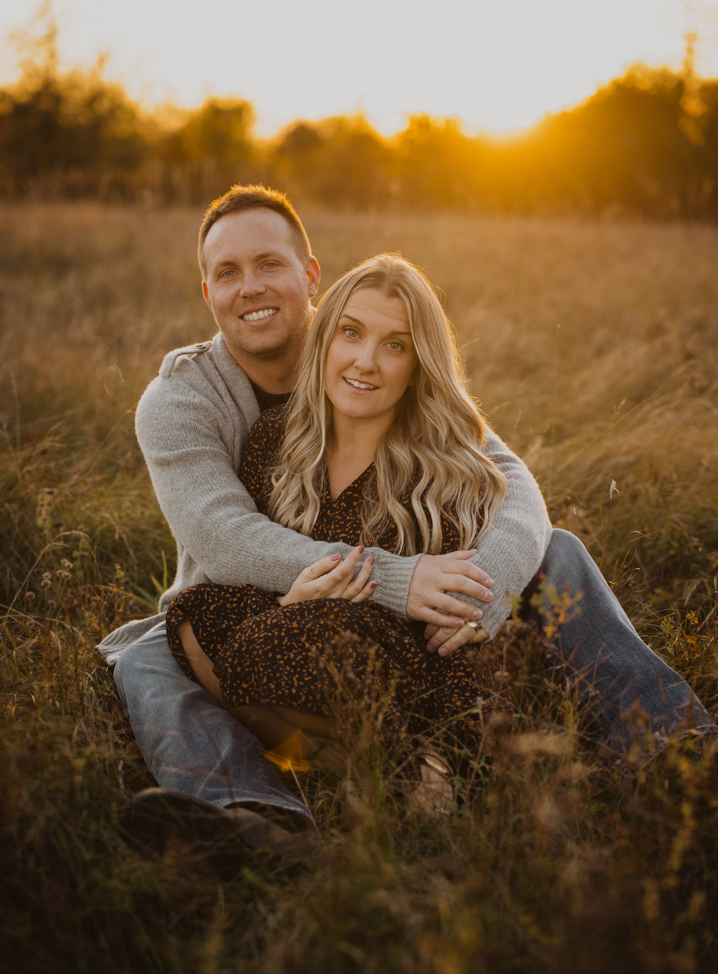 A romantic photo of a couple sitting together in a sunlit field at sunset, embracing and enjoying a peaceful moment.