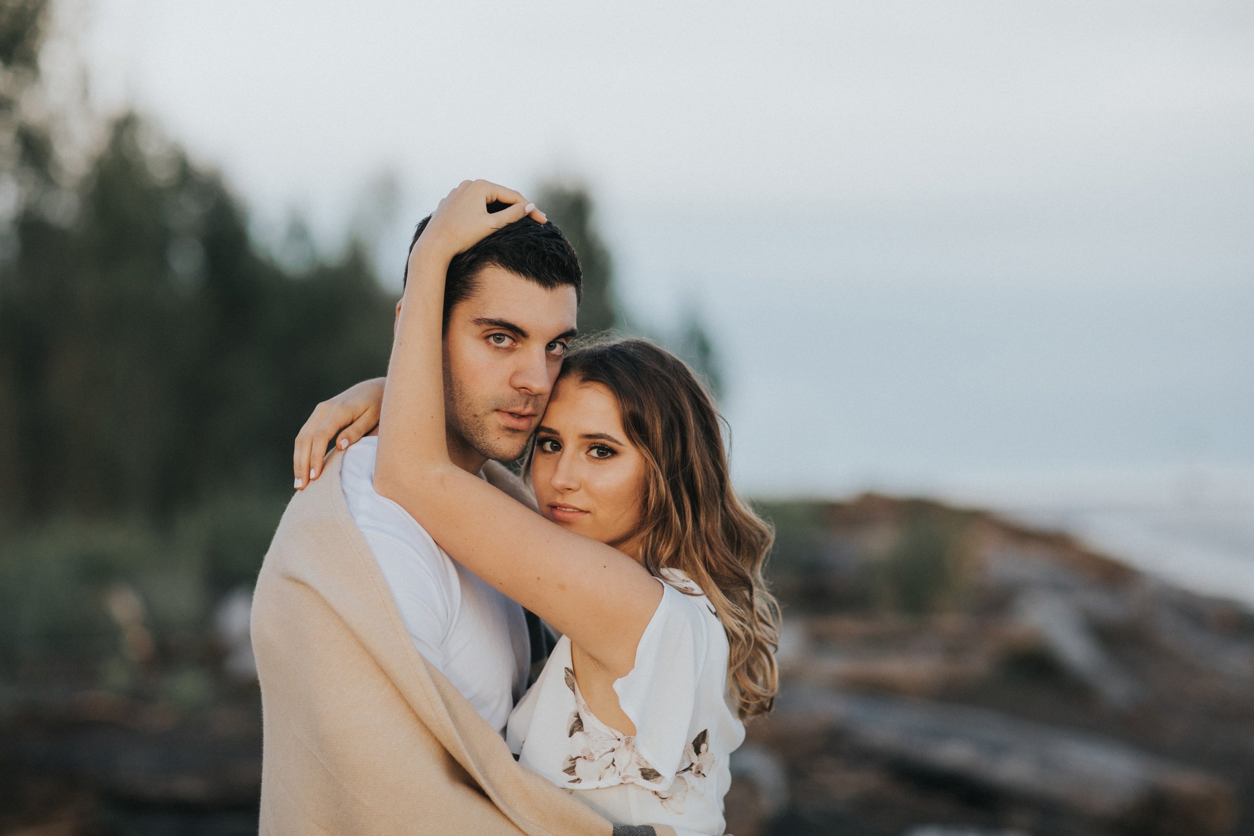 A couple wrapped in a blanket shares a tender moment on the beach at sunset, with the cool breeze and ocean in the background.