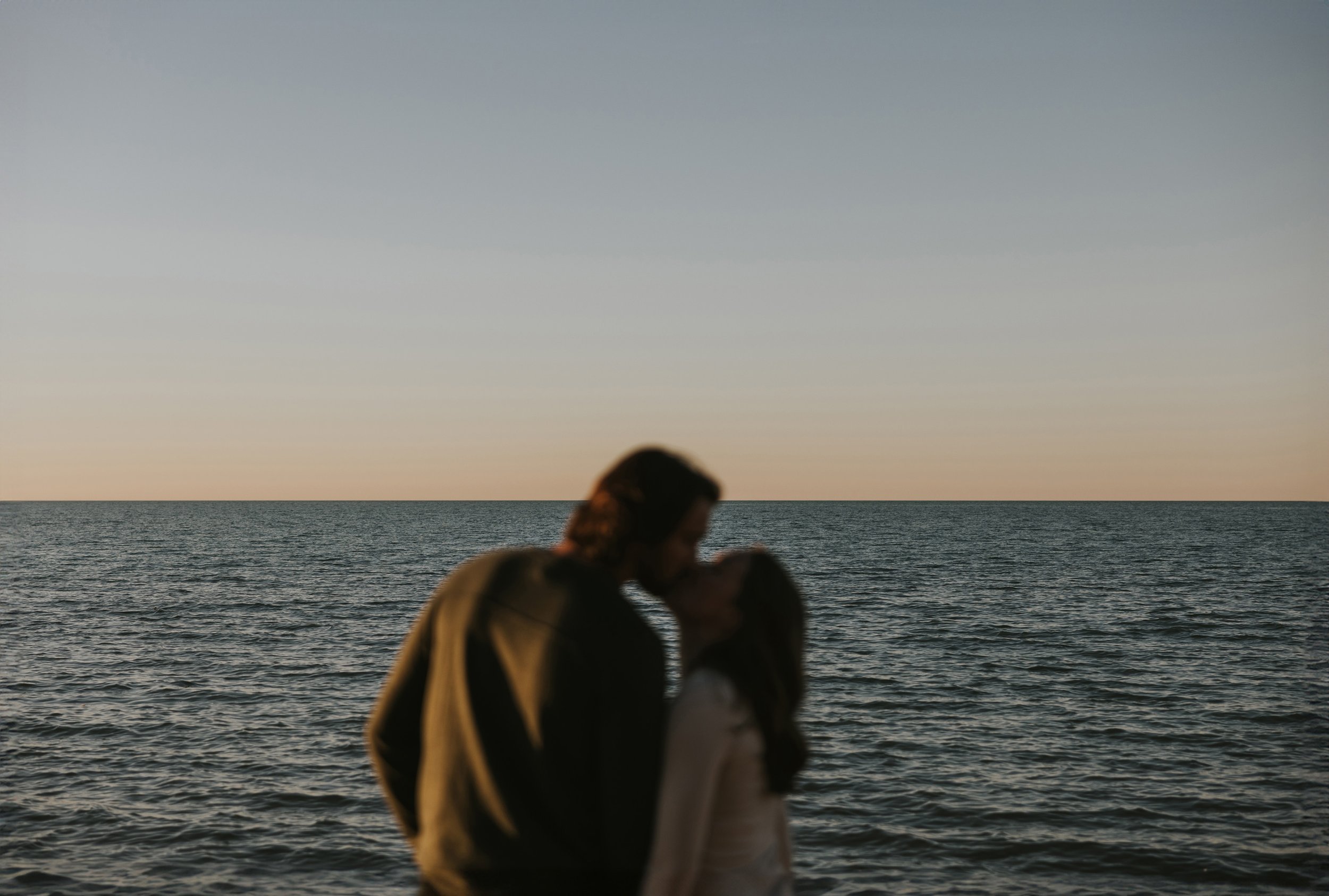 A couple shares a kiss in silhouette against the backdrop of a beach sunset, the calm ocean and warm colours enhancing the romantic mood.