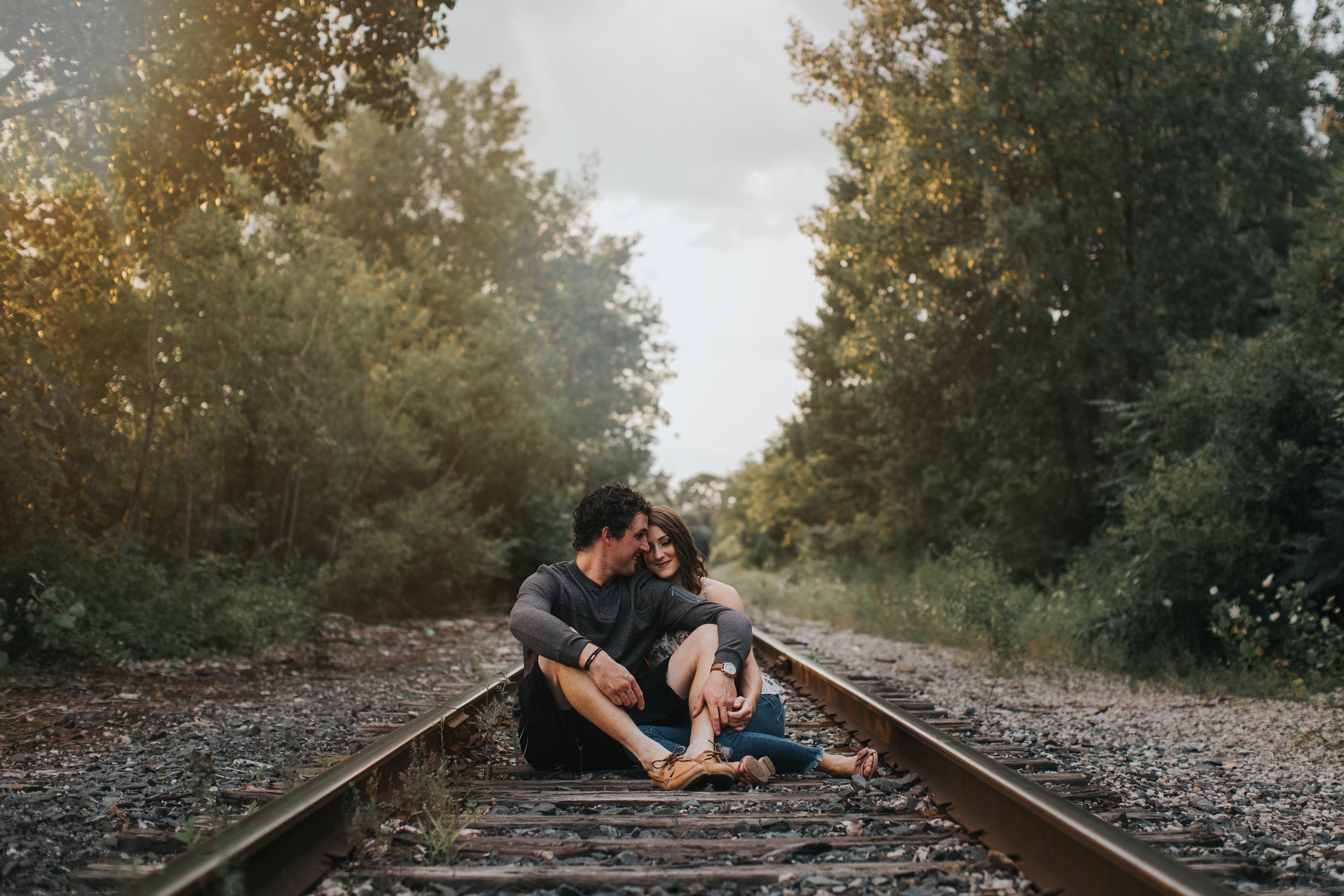 A couple sitting together on railroad tracks, surrounded by lush green trees. They are leaning in close, with the man resting his head on the woman's forehead as they share an intimate moment. The warm light of the setting sun filters through the tre