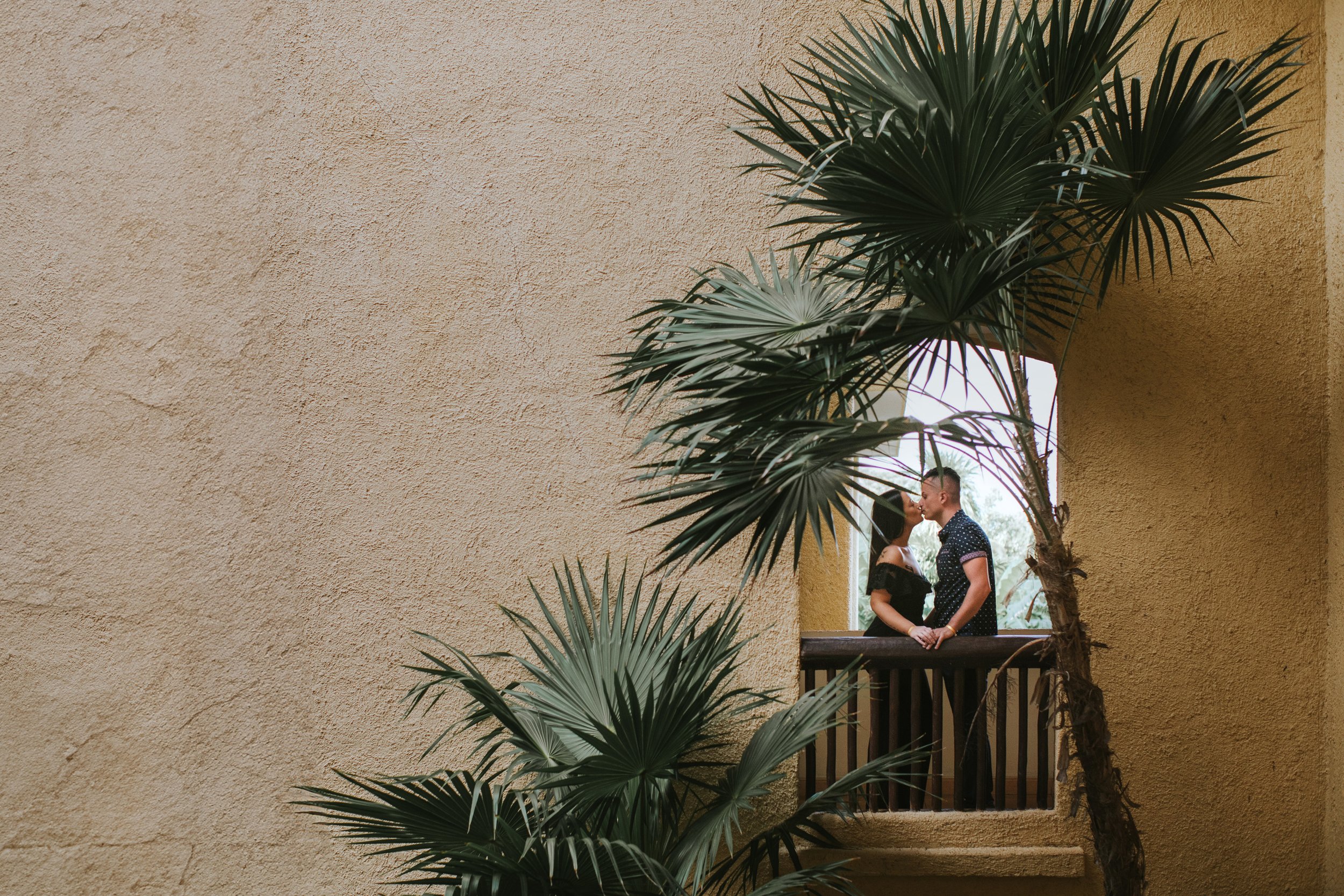 A couple shares a tender kiss on a tropical resort balcony, surrounded by palm trees, capturing the beauty and romance of their engagement.