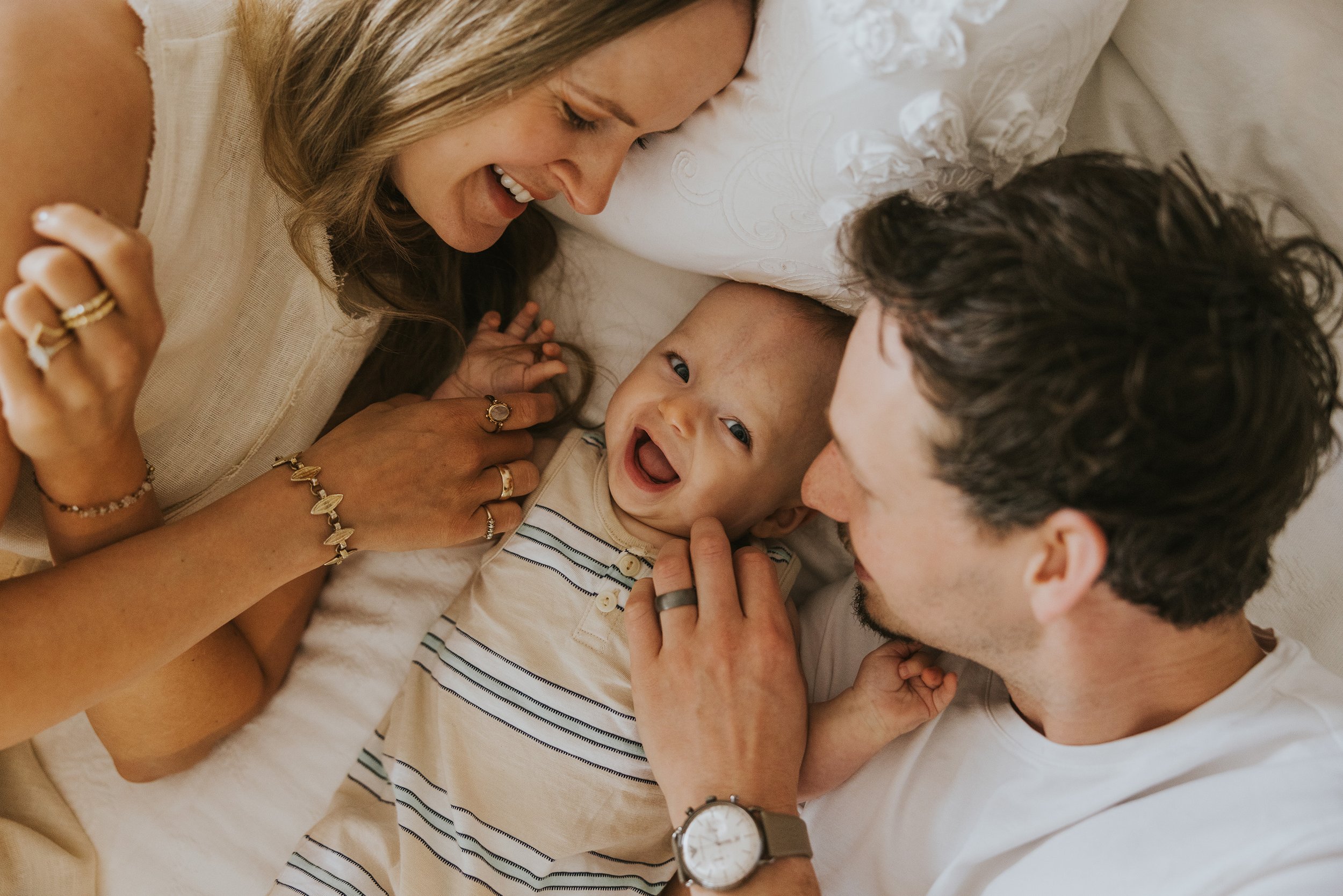 Parents lying on a bed, joyfully playing with their smiling baby, capturing a loving and playful family moment.